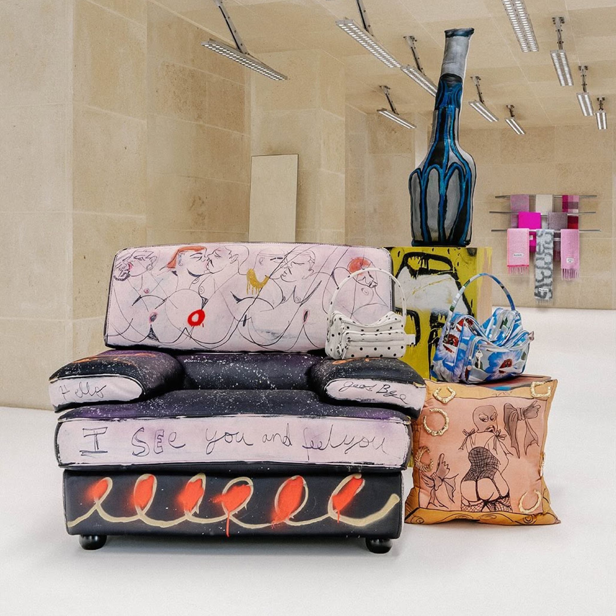Artistic view representing the Metamorphosis trend, featuring an upholstered armchair covered in expressive figurative artwork and text, paired with eclectic accessories like patterned handbags and a sculptural bottle. The mix of reimagined objects highlights how everyday items gain new meaning through creative transformation.