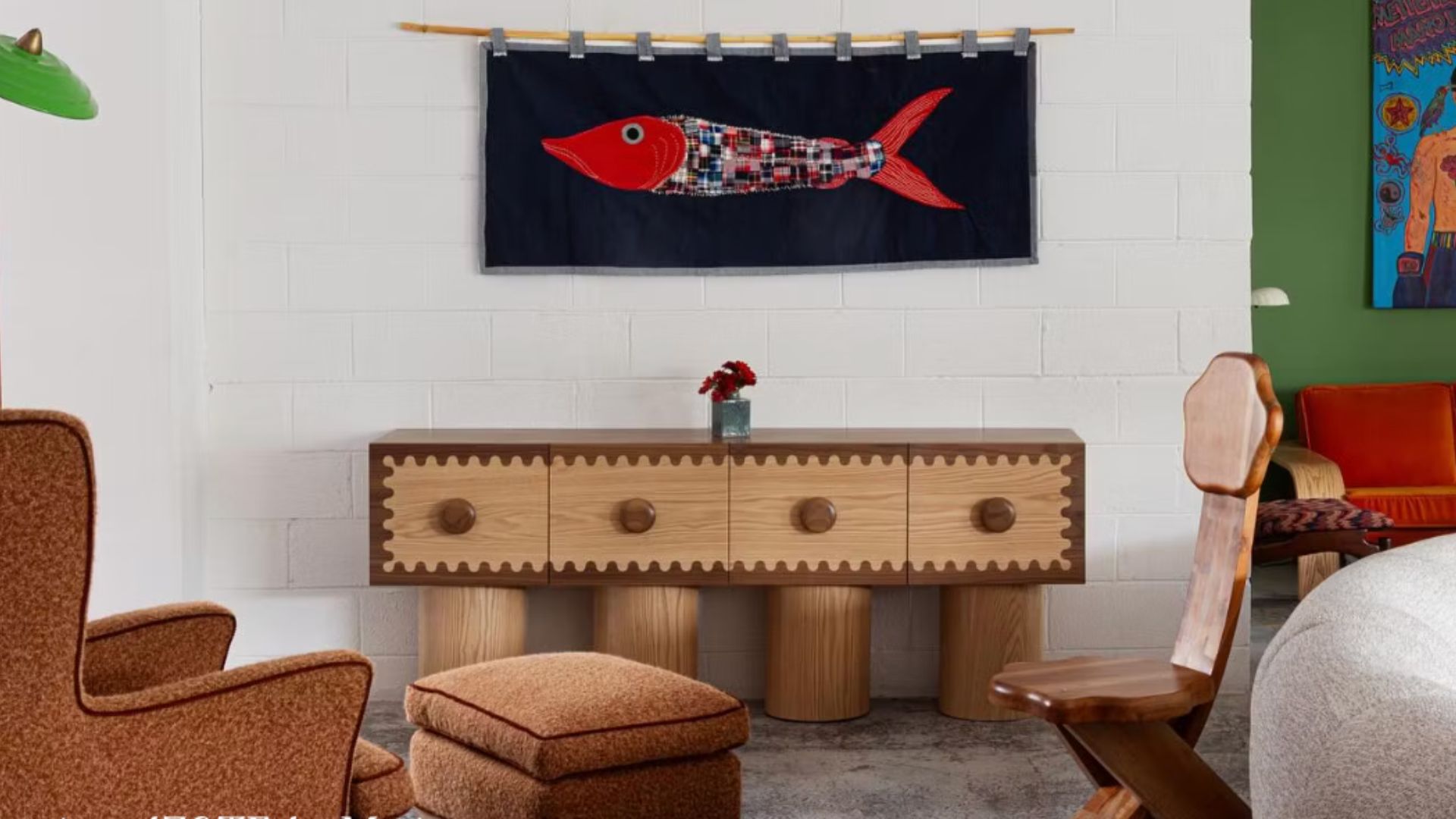 Eclectic interior scene representing the Neo-Folklore trend, featuring a sculptural wooden sideboard with playful circular knobs and zigzag detailing. Above it hangs a textile artwork of a red fish, evoking folk symbolism through patchwork and handcrafted textures. Surrounding furniture and vibrant accents blend retro charm with artisanal warmth, celebrating local identity and personal storytelling in contemporary design.