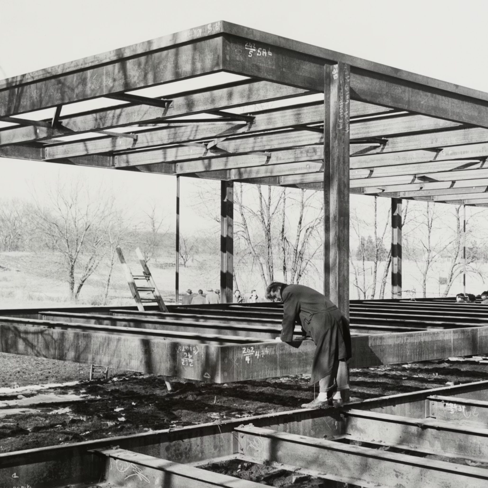 A worker stands atop the exposed steel frame of the Farnsworth House structure, marking measurements across the beams as the skeletal form rises against a backdrop of bare winter trees. The scene captures the early construction phase of the Farnsworth House, where its delicate proportions and minimalist design first took shape. The raw steel, still covered in chalk notations, reveals the precision and discipline behind Mies van der Rohe’s vision, long before the glass enclosure transformed it into a place of retreat.