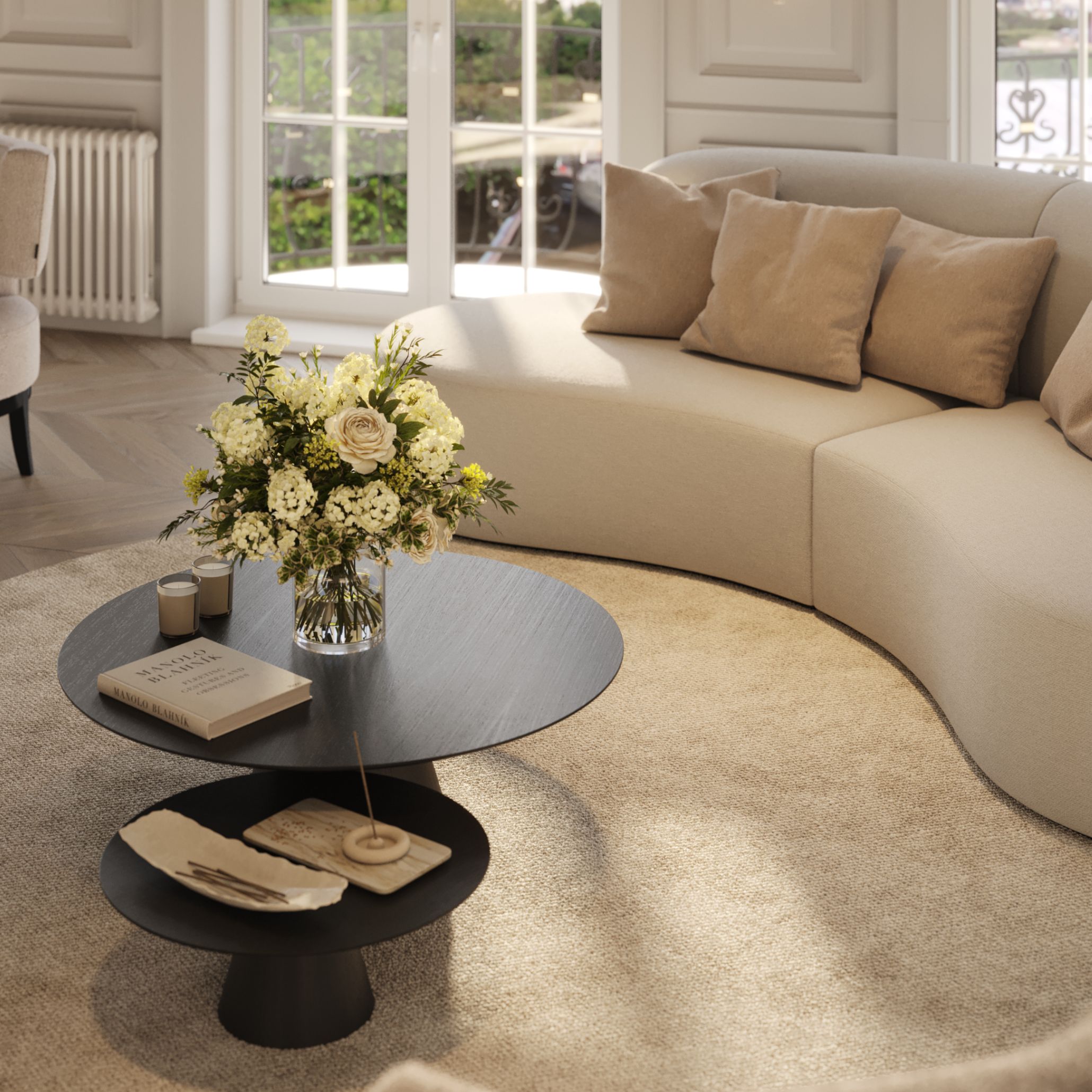 A refined living room design featuring the Cotton Sofa in warm beige upholstery, its curved silhouette embracing a central arrangement of Sahara luxury coffee tables. The scene highlights contemporary furniture design through soft forms, layered texture, and modern furniture details: a floral arrangement, sculptural candles, and a design book resting on the table’s dark surface. Natural light pours in through tall French windows, illuminating the gentle palette and enhancing the tactile materials that define this serene, modern interior.