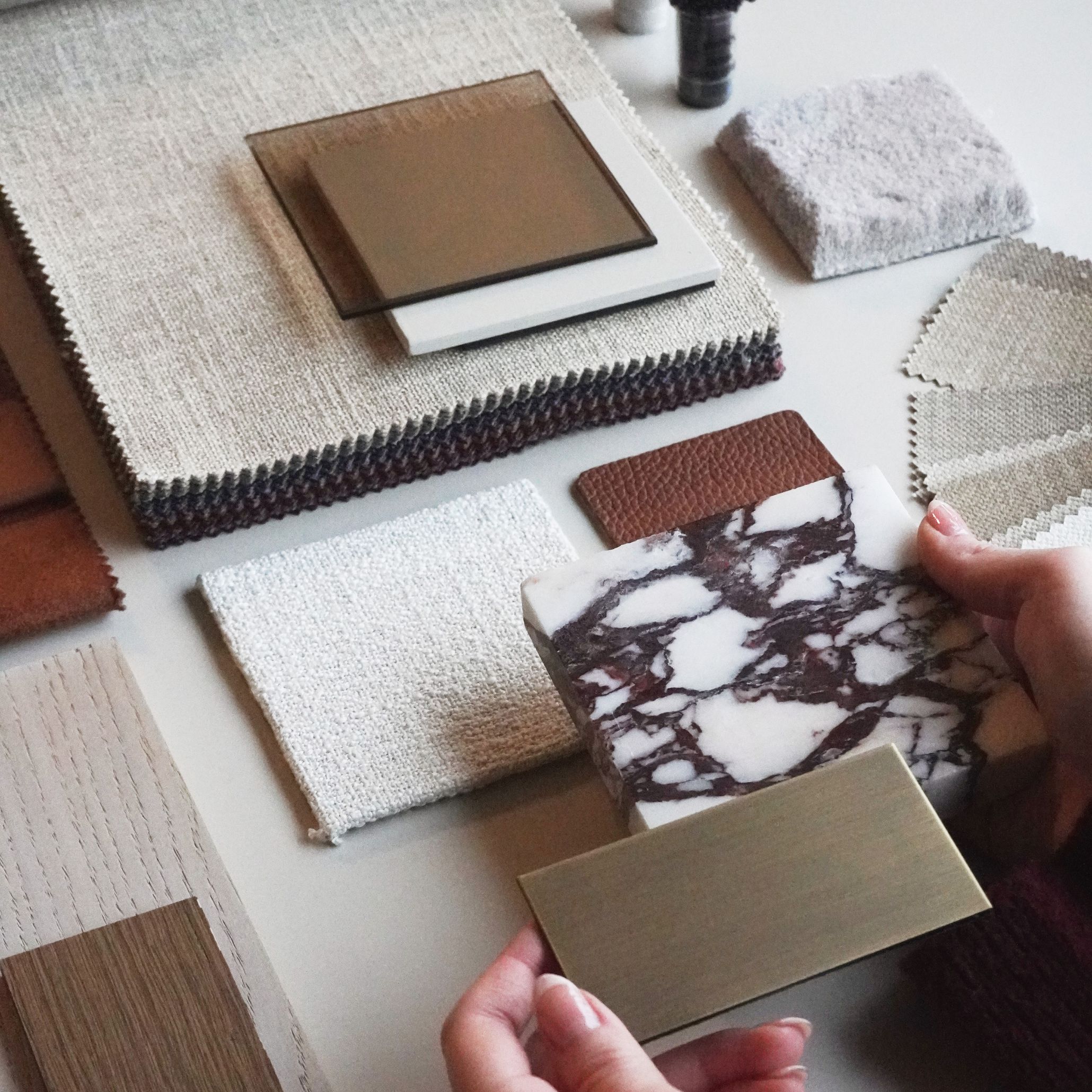 A curated selection of materials showcasing marble, textured fabrics, wood veneers, and metal finishes arranged as interior design inspiration. The composition highlights the tactile beauty central to contemporary furniture design, with hands exploring design details that emphasize contrast, craftsmanship, and the sensory qualities of each material.