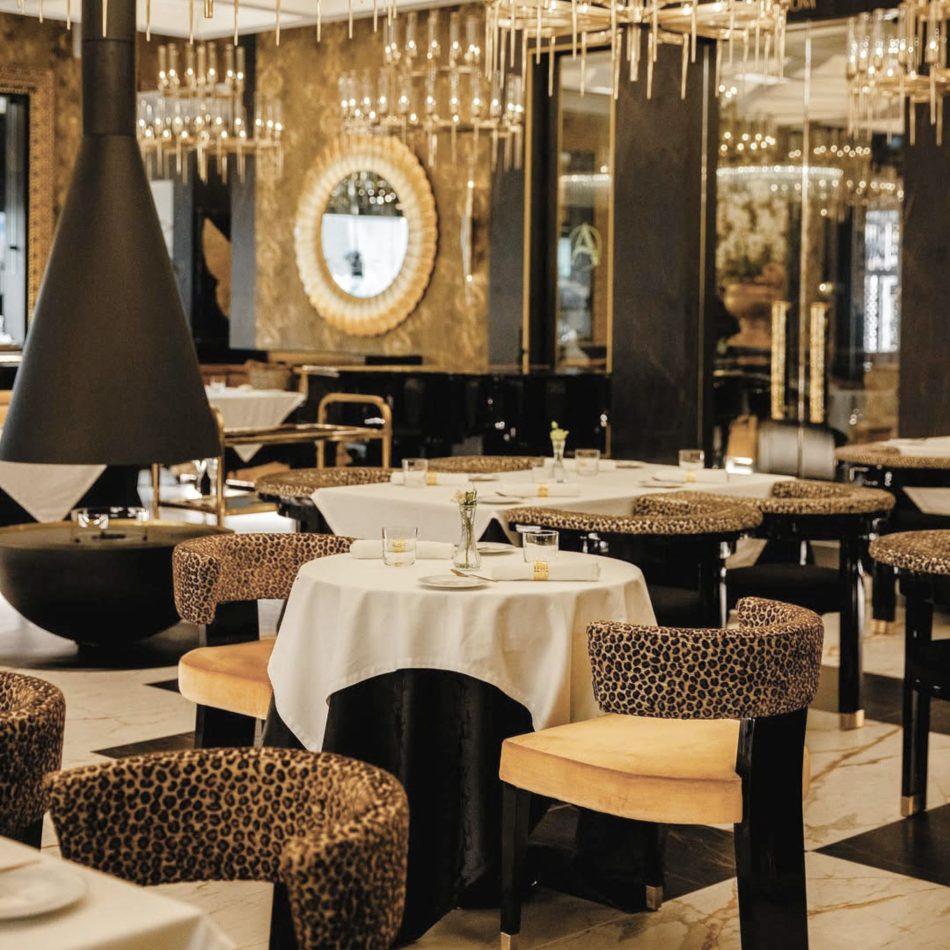An elegant dining room where sunlight highlights round tables dressed in layered linens, surrounded by the bold Her II Chair in patterned upholstery. Gold accents, marble flooring, and a sculptural fireplace enrich the atmosphere, blending restaurant dining chairs, refined luxury furniture, subtle design details, warm gold touches, and a sense of hospitality furniture sophistication.