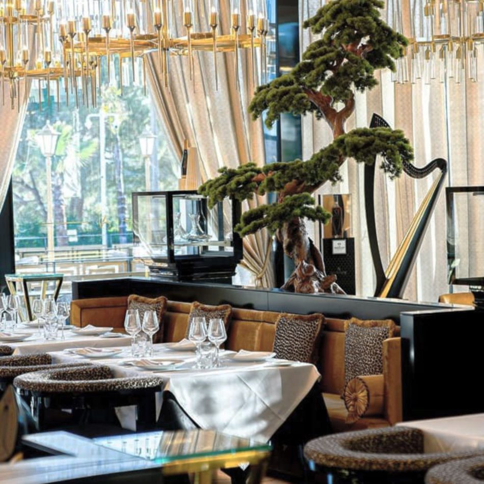 A luminous restaurant interior centered around a sculptural bonsai tree, with tables set beneath cascading glass chandeliers and framed by soft drapery. The Kensington Chair introduce rich textures and statement silhouettes, complemented by restaurant furniture, curated hospitality designs, elegant gold-plated-brass accents, and thoughtful design details that elevate the dining experience.