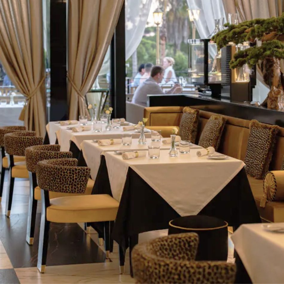 A refined dining space featuring patterned restaurant chairs, a long upholstered banquette, and tables arranged with delicate glassware and single‑stem florals. Sunlight filters through tall curtains toward the terrace, enhancing the harmony between modern restaurant dining chairs, elegant restaurant decor ideas, layered design details, inviting hospitality furniture, and a warm, luxurious ambiance.