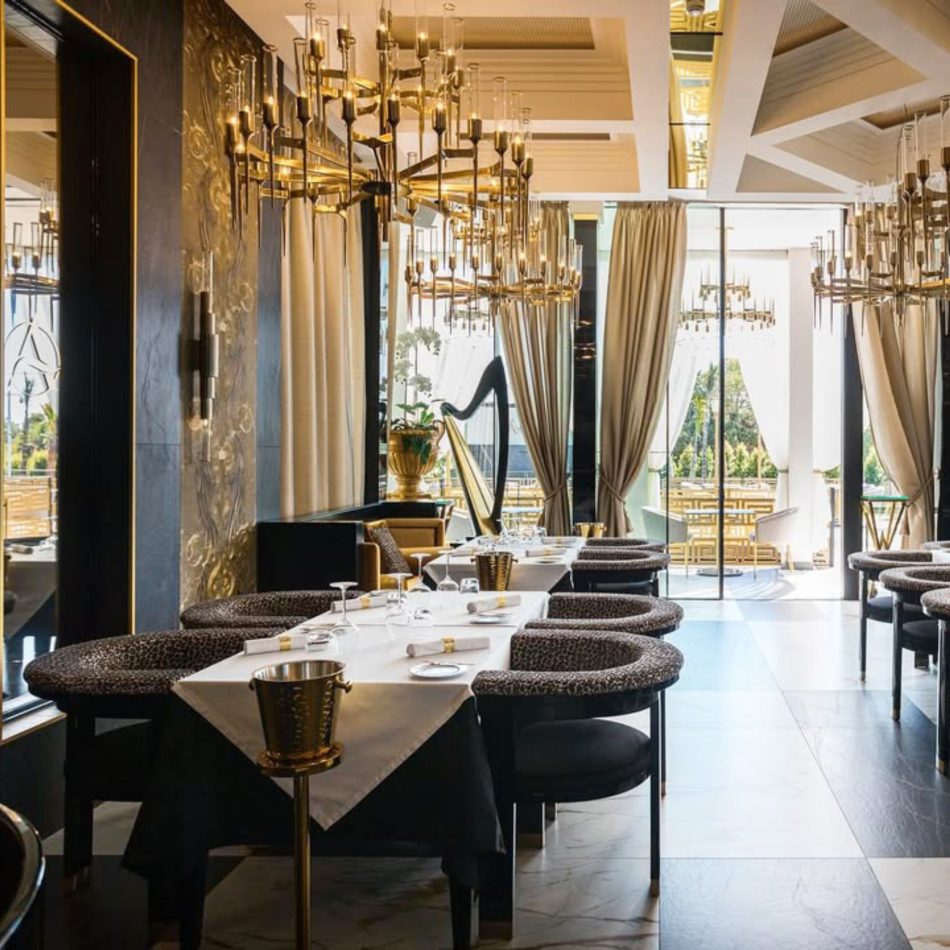 An opulent restaurant interior featuring gold‑toned chandeliers, patterned seating, and tables arranged for an elevated dining experience, framed by tall curtains and a harp near the window. The warm glow highlights modern bar furniture, refined restaurant table, sculptural bar furniture, thoughtful design details, and the inviting presence of luxury dining room furniture.