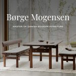 A minimalist dining setting features a dark solid‑wood table paired with two matching benches upholstered in soft grey cushions, arranged on a light woven rug before large floor‑to‑ceiling windows that open to a calm courtyard with pebbles and climbing greenery, expressing the balanced proportions, crafted simplicity, and functional elegance associated with Danish Modern furniture and Børge Mogensen’s design philosophy.