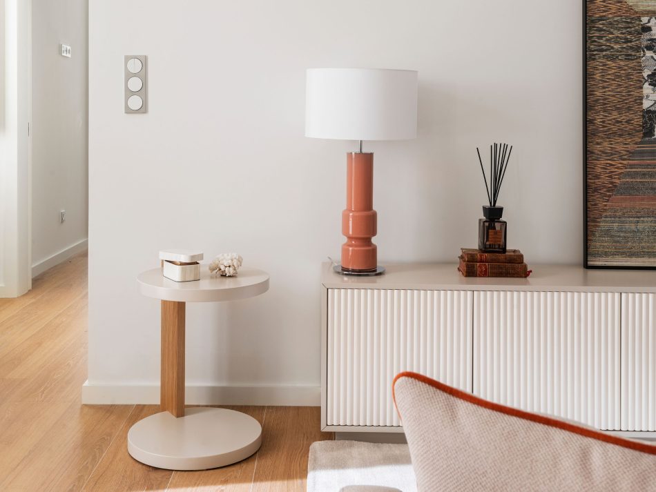 A close, composed corner of the living room where the Club Side Table holds a small decorative piece beside the Ryan TV Cabinet, whose vertical rhythm and warm tones anchor the space. Soft natural light and earthy accents create a tranquil setting enriched by modern furniture, warm home decor inspiration, and the understated elegance of crafted materials.