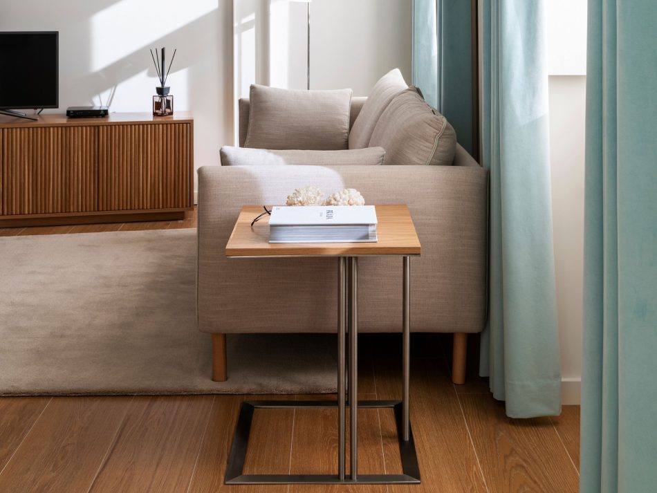 A quiet living room vignette where the Louise Side Table rests beside a soft upholstered sofa, its simple form adding a gentle functional note to the guest residence. Warm wood flooring, diffused natural light, and a calm palette create an atmosphere of modern furniture, understated living room design, and the intimate elegance that welcomes every guest.