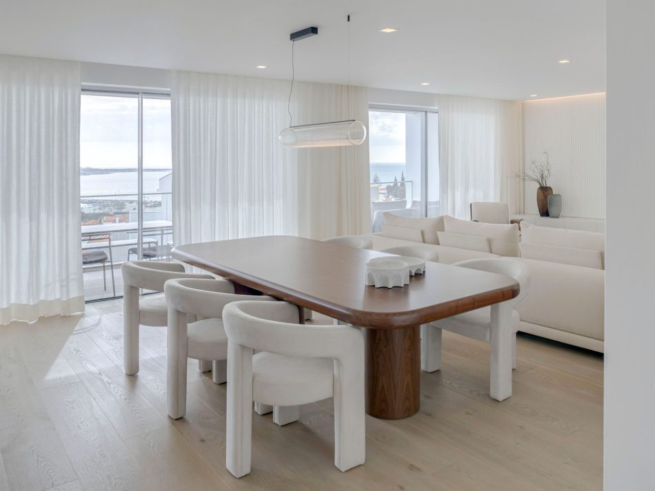A bright open‑space dining area where natural light flows through large windows, illuminating the soft textures and organic materials that define the room. At the center, the Natur Dining Table anchors the space with its sculptural wooden form, creating a warm transition toward the adjoining living area. The atmosphere blends luxury dining room furniture with subtle touches of modern furniture and a sense of interior design inspiration shaped by simplicity and light.