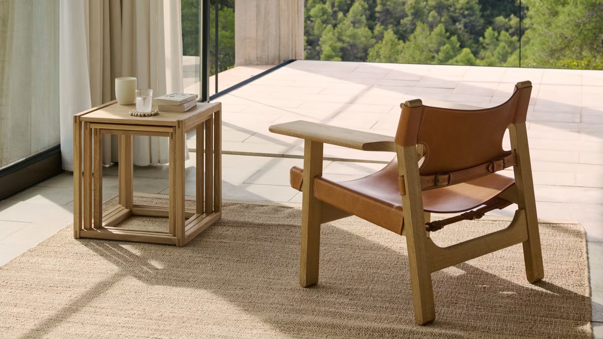 A Spanish Chair in natural oak with wide leather straps sits beside the BM375 Nesting Tables arranged on a textured rug, framed by a calm indoor‑outdoor setting that highlights luxury living room furniture, solid wood craftsmanship, and modern furniture design.