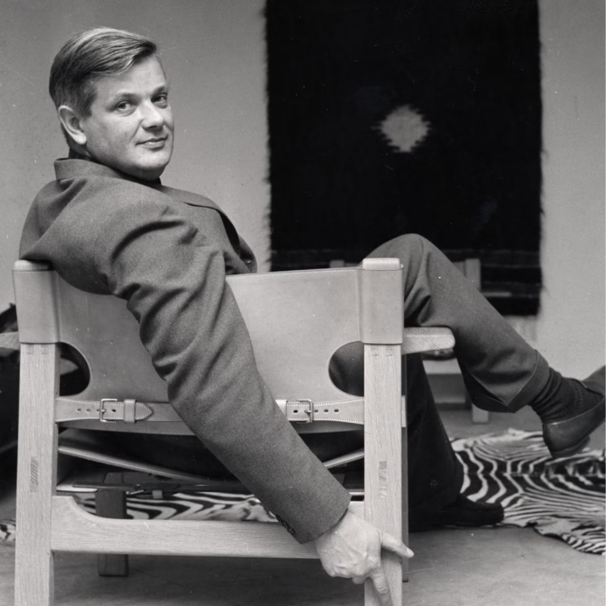 A black‑and‑white portrait of Børge Mogensen seated in a the Spanish Chair chair with leather straps, set against a patterned rug and textile wall hanging, capturing the material honesty and functional clarity of mahogany furniture, the restrained elegance of modern furniture, and the timeless craftsmanship associated with furniture luxury design.