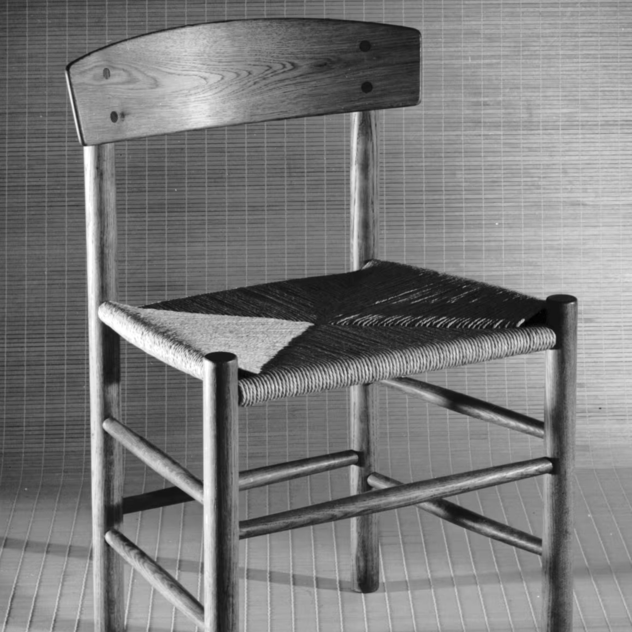 A J39 Chair with its simple solid‑wood frame and tightly woven paper‑cord seat stands against a textured backdrop, reflecting Mogensen’s commitment to functional clarity through wood texture, finest wood craftsmanship, and the enduring principles of contemporary furniture design.