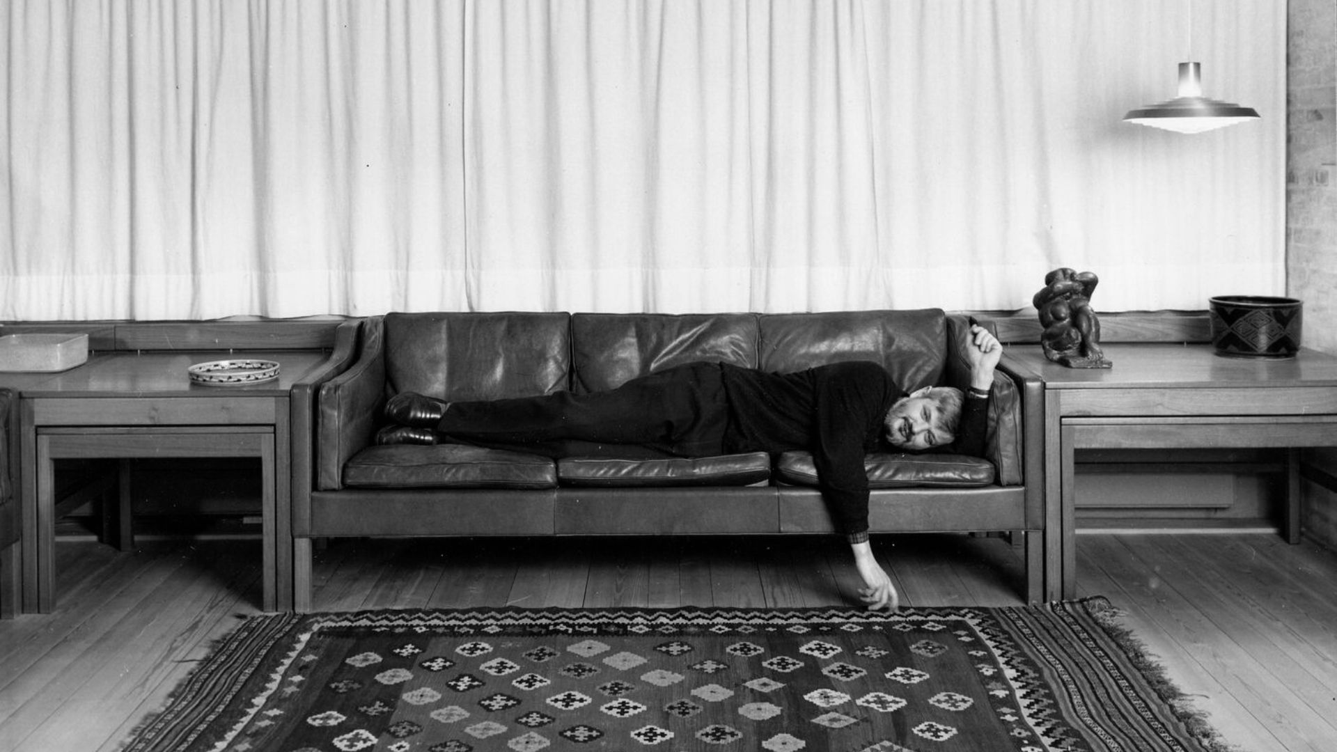 Børge Mogensen rests on a leather sofa in a quiet, minimalist living room with wooden side tables, a patterned rug, and sculptural decor, embodying his human‑centred approach to comfort through leather upholstery, living room furniture, and the honest warmth of solid wood craftsmanship.