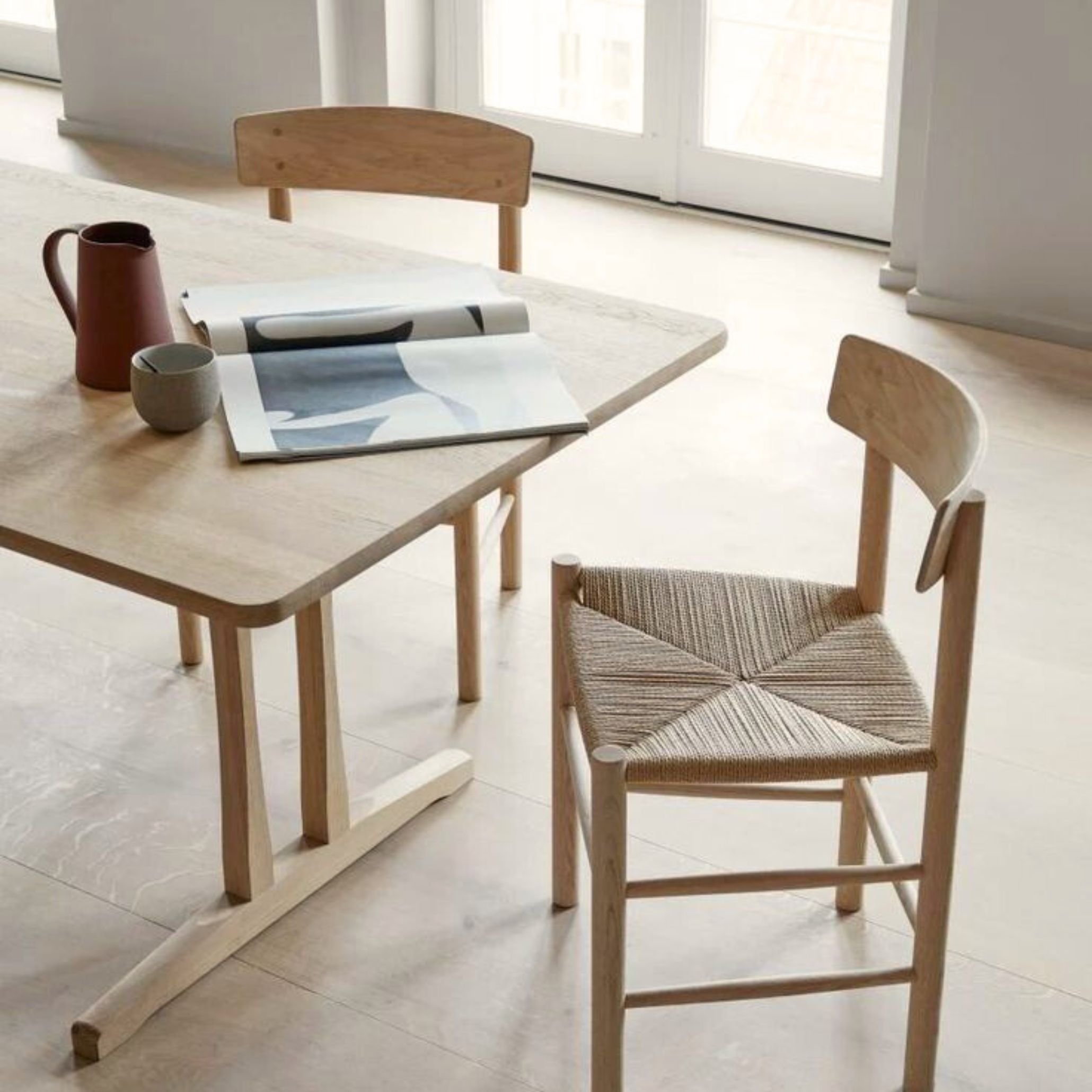 A J39 Mogensen Chair in light wood with a woven paper‑cord seat stands beside a rounded‑edge dining table in a bright, minimalist room, expressing the Danish Modern clarity of finest wood craftsmanship, the functional simplicity of a modern dining table, and the enduring appeal of contemporary furniture.