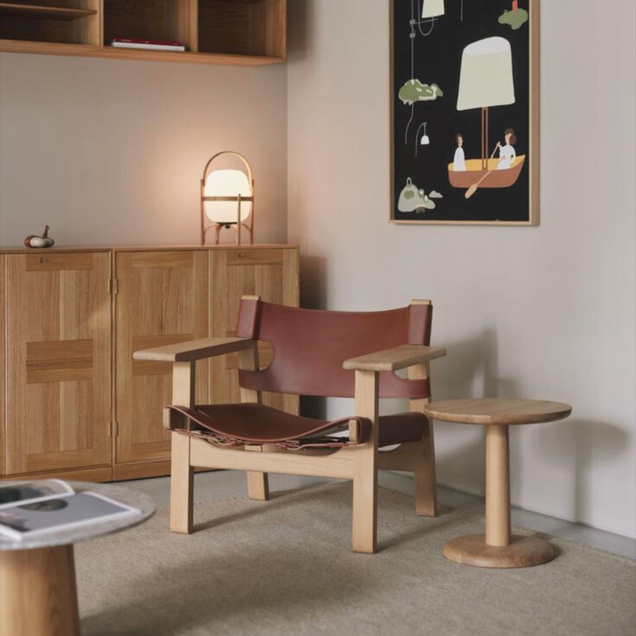 A Spanish Chair in natural oak with wide brown leather straps sits beside a round wooden side table and a light‑wood cabinet with a sculptural lamp, creating a warm, minimalist interior that reflects leather furniture craftsmanship, the quiet refinement of luxury home furniture, and the timeless clarity of contemporary furniture design.