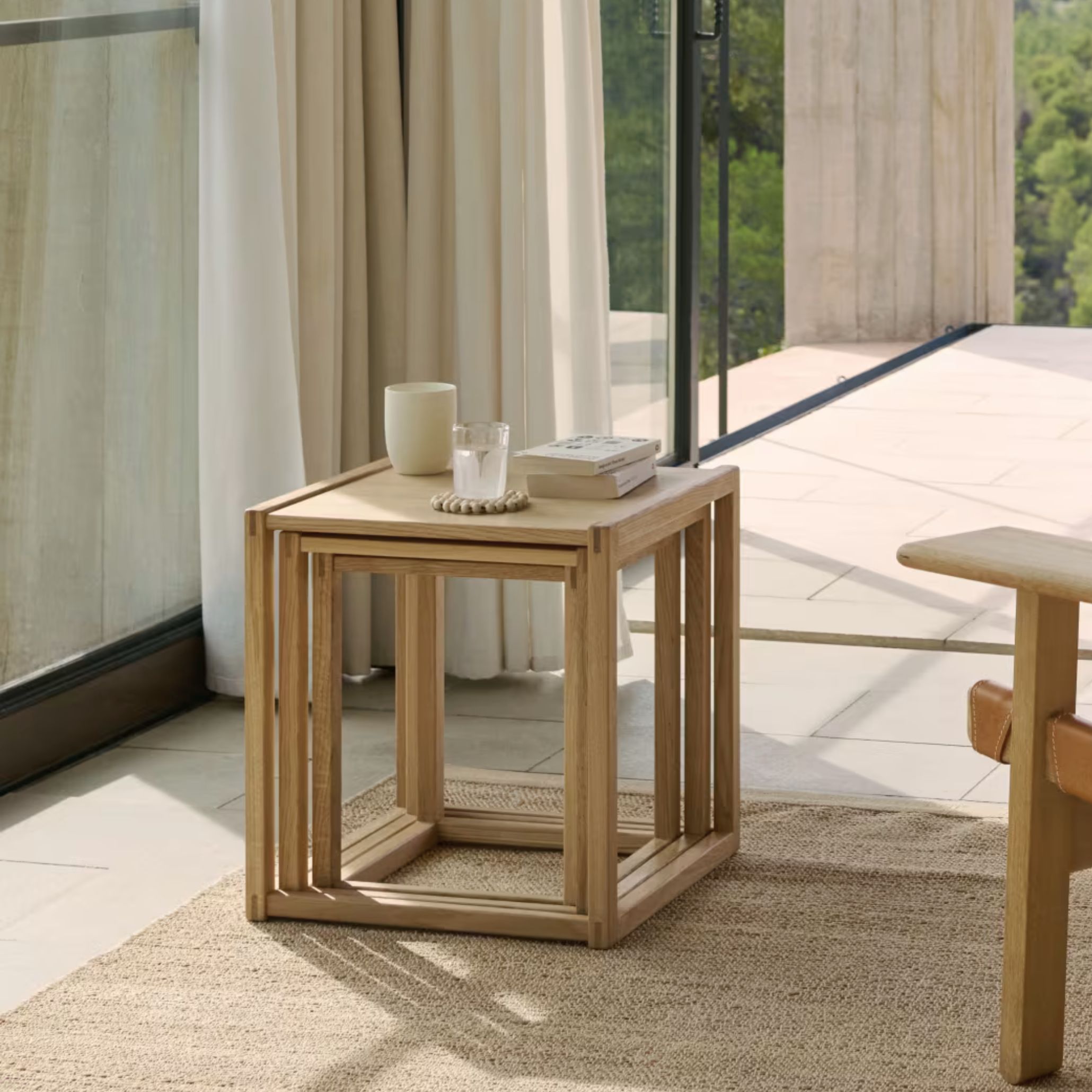 The BM375 Nesting Tables in light solid wood are arranged on a woven rug beside floor‑length curtains, styled with a ceramic cup, candle holder, and books, expressing the quiet functionality of modern side tables, the natural warmth of wood texture, and the refined simplicity of contemporary furniture design.