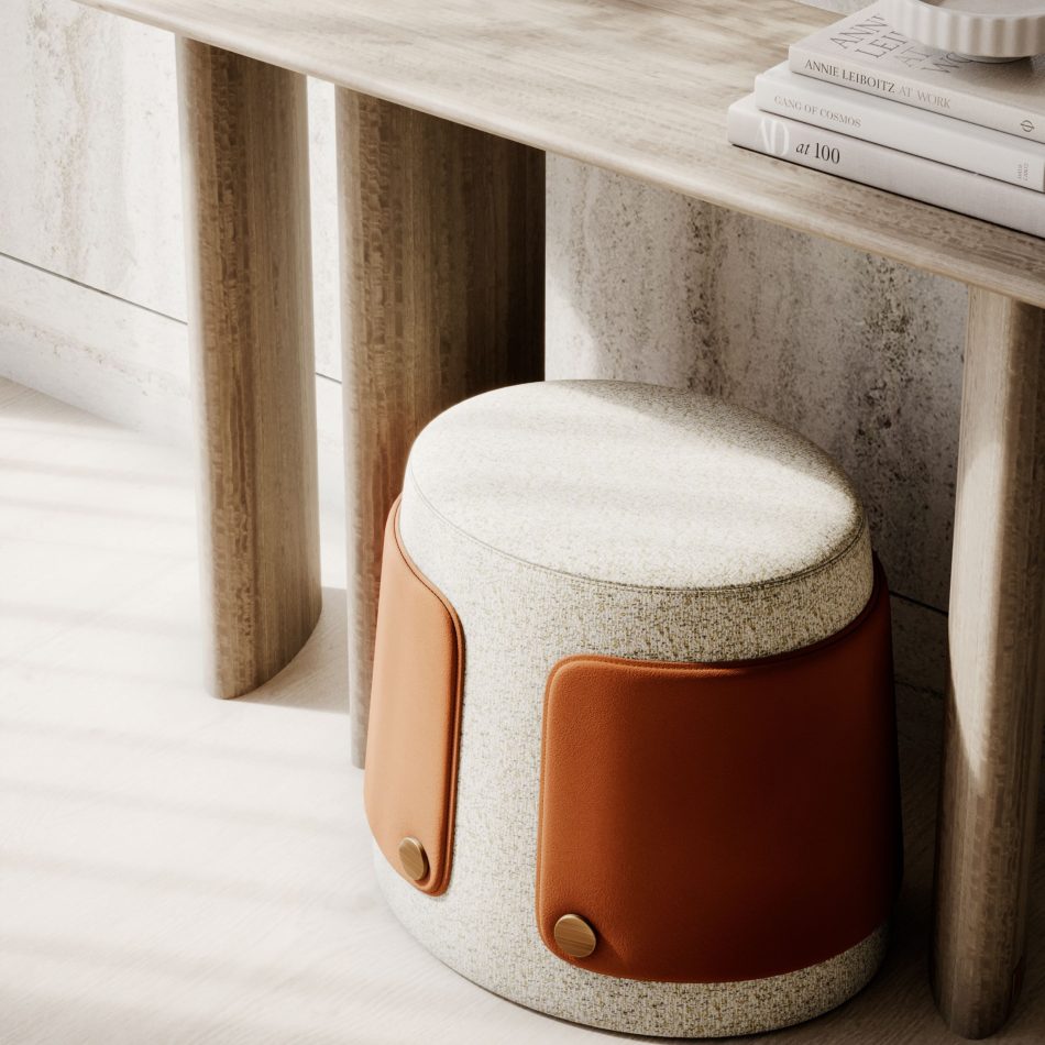 The Dolomites Stool sits beneath a cylindrical‑leg console, illuminated by natural light that casts soft shadows across the textured wall and wooden surfaces. Its upholstery and leather panels fastened with brass buttons introduce a tactile richness that feels both grounded and delicate, echoing landscapes shaped slowly over time. The books, warm tones, and minimalist styling create an atmosphere of quiet contemplation, positioning the stool as a accent within modern furniture and refined living room architecture.