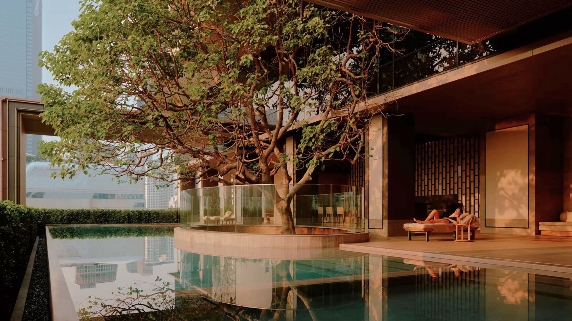 A serene wellness lounge at Aman Nai Lert Bangkok, where contemporary architecture blends seamlessly with nature. A reflective pool sits beside an open relaxation area furnished with minimalist living room furniture and warm lighting, while a large tree is integrated into the design to honour cultural rituals of rest and renewal. The tranquil atmosphere reflects wellness architecture supported by thoughtfully curated luxury home furniture that nurtures both body and mind.