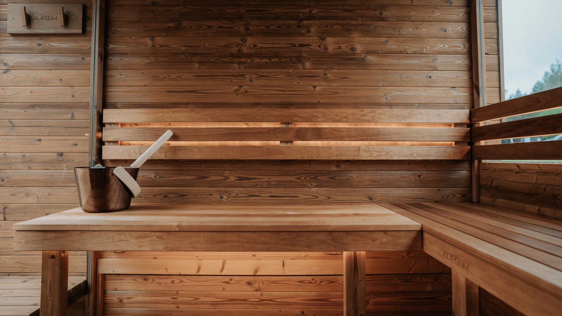 A warm Nordic sauna interior crafted entirely from smooth wood, featuring corner benches and a central bucket for steam rituals. Natural light filters through a small window, enhancing the calming atmosphere and the simplicity of solid wood design. The space reflects traditional sauna culture, where heat, rest, and community come together, supported by subtle touches of luxury home furniture and the purity of handmade furniture craftsmanship.