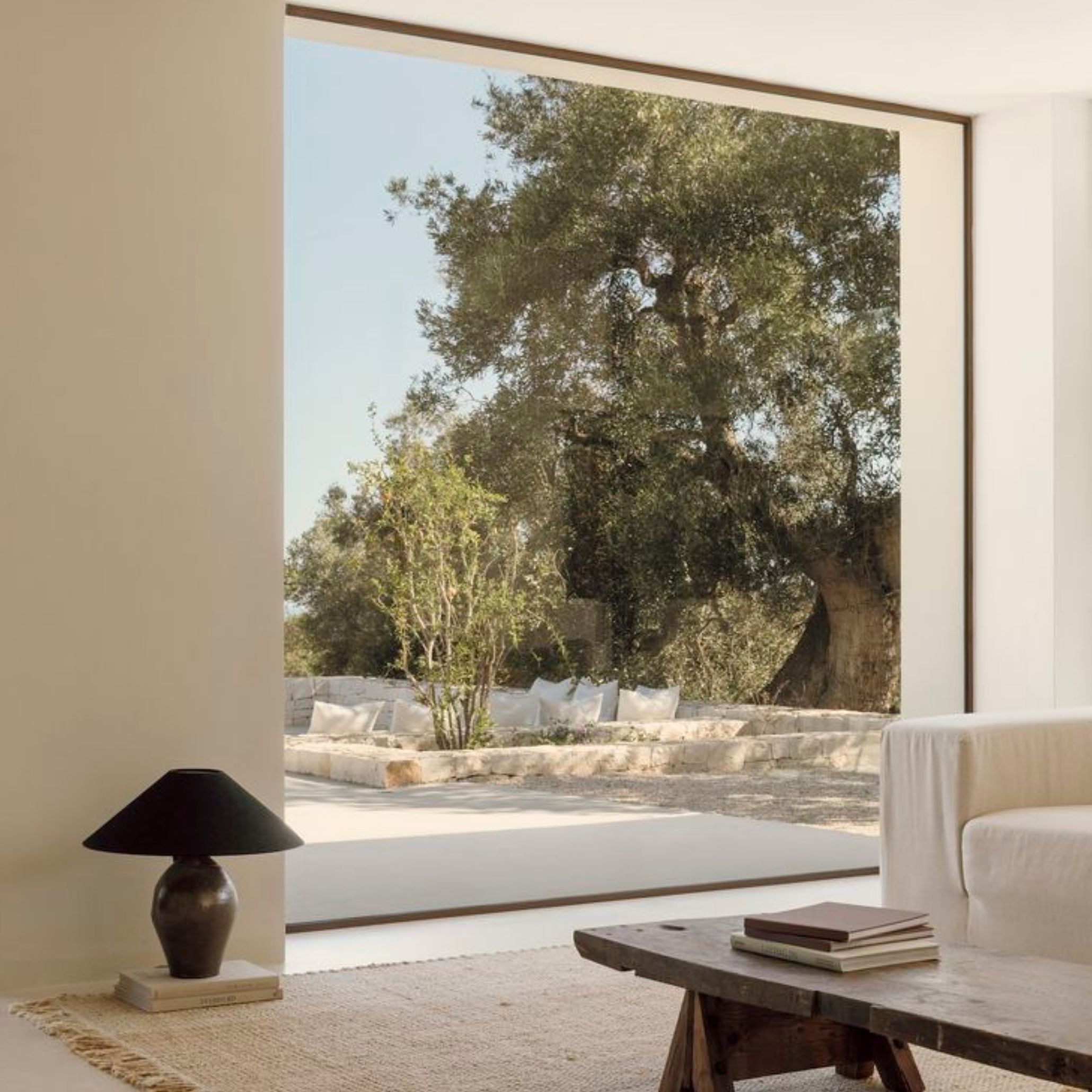 A calm indoor‑outdoor living space where natural light pours through expansive windows, illuminating a white sofa and rustic wooden coffee table. The seamless connection to the stone patio and surrounding greenery reflects wellness‑driven interiors shaped by natural materials, refined interior design, and the gentle warmth of organic textures that support light and airflow throughout the home.