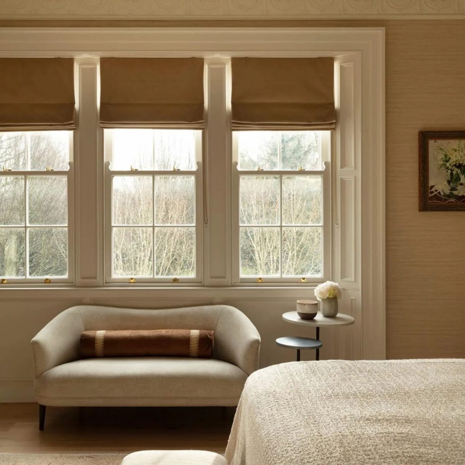 A serene master‑suite sitting area with a soft upholstered loveseat placed before three tall windows dressed in warm Roman shades, filling the room with natural light. A round side table with delicate décor complements the calm palette, reflecting luxury master bedroom furniture, subtle bedroom architecture, and refined design details that echo the project’s timeless character and sophisticated bedroom furniture aesthetic.