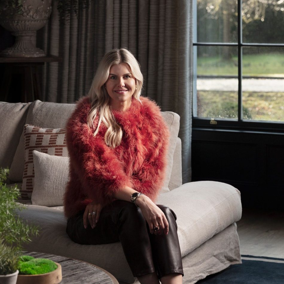 The designer sits on a grey sofa in a serene living corner framed by tall curtains and a garden view, surrounded by patterned pillows, greenery, and a round coffee table. Soft natural light enhances the warm textures and calm palette, reflecting luxury home furniture, gentle interior design inspiration, refined living room architecture, and the understated elegance of modern furniture within the residence.
