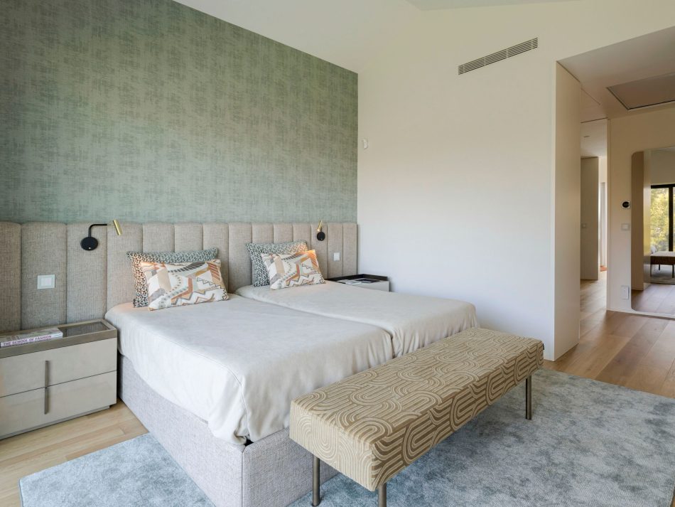 A bright master suite with twin beds sharing an upholstered headboard, complemented by a patterned bench that anchors the room. The Slender Mirror in the hallway subtly expands the space and reinforces the calm atmosphere. Soft materials and warm tones reflect luxury master bedroom furniture, elegant bedroom architecture, gentle wood texture, and curated sophisticated bedroom furniture.