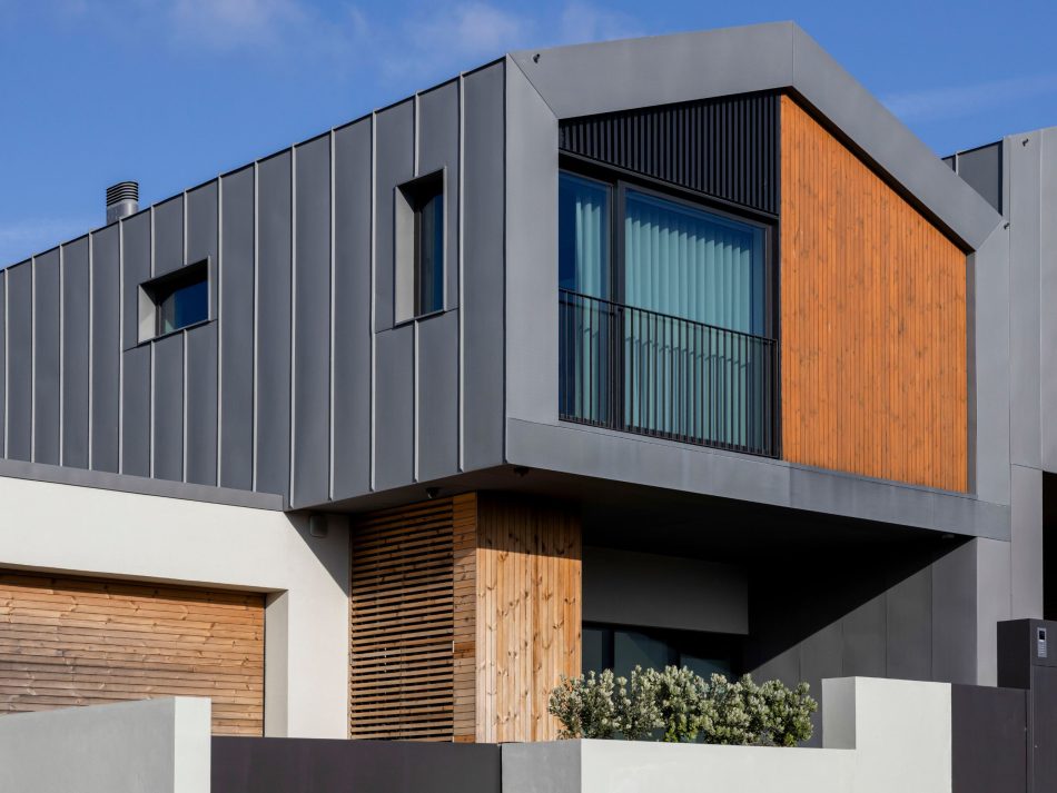 A contemporary two‑story home featuring a minimalist architectural design with clean geometric lines. The exterior combines dark metal cladding, smooth light‑toned concrete, and vertical natural‑wood paneling for a layered, modern look. The upper floor includes expansive glass windows with vertical blinds and a slim balcony framed by a black metal railing. The lower level showcases a wide wooden garage door, a section of vertical wooden slats, and a small landscaped area with neatly trimmed shrubs, emphasizing the home’s warm‑modern aesthetic.