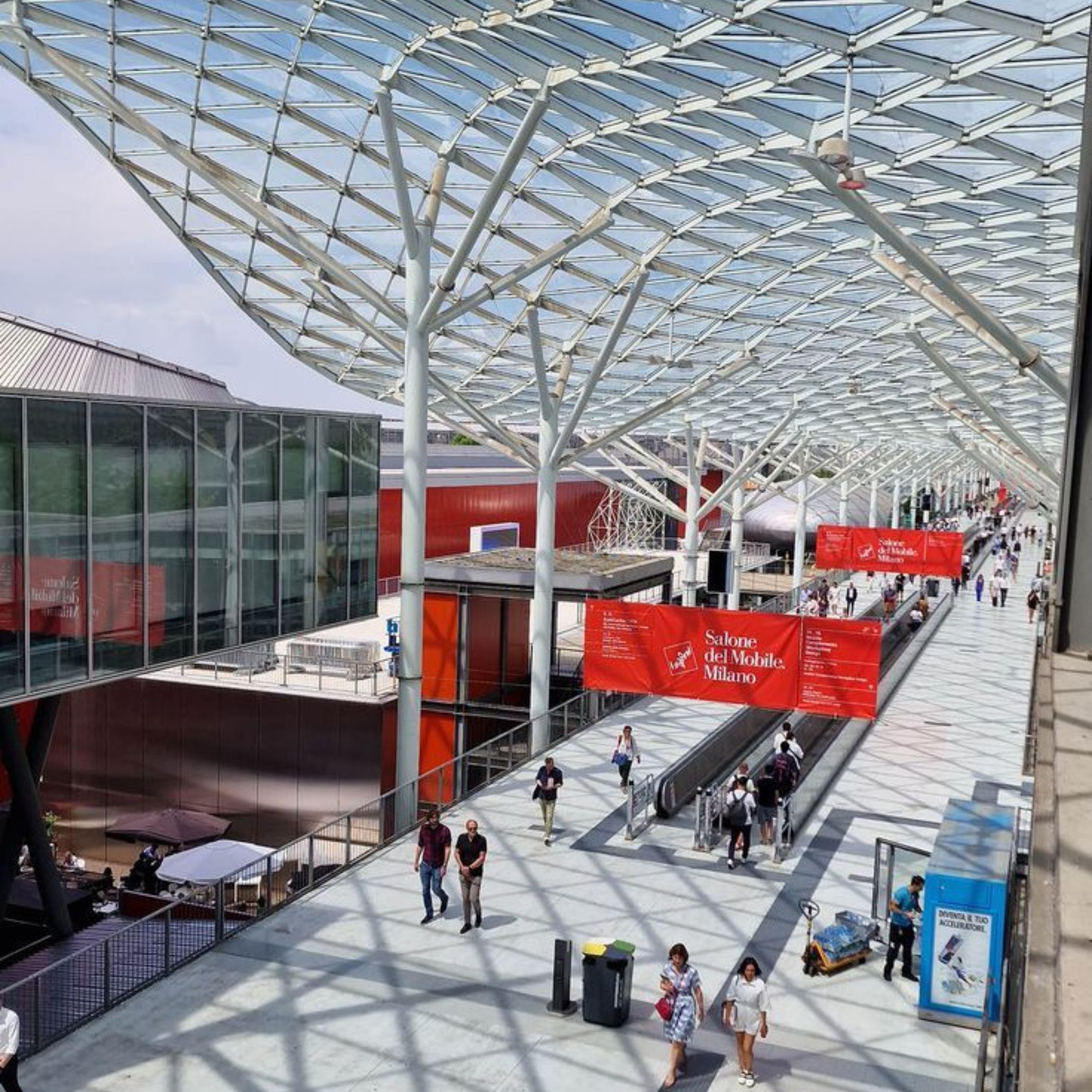Visitors move along the glass‑covered walkway at Fiera Milano, framed by geometric steel lines and red Salone del Mobile banners that signal the start of Design Week. The architectural scale and flow of people reflect a global gathering shaped by modern furniture innovation, where material honesty and longevity guide new directions in luxury furniture. The scene captures the energy of Milan as it becomes a hub for interior design inspiration, grounded in the theme A Matter of Salone.
