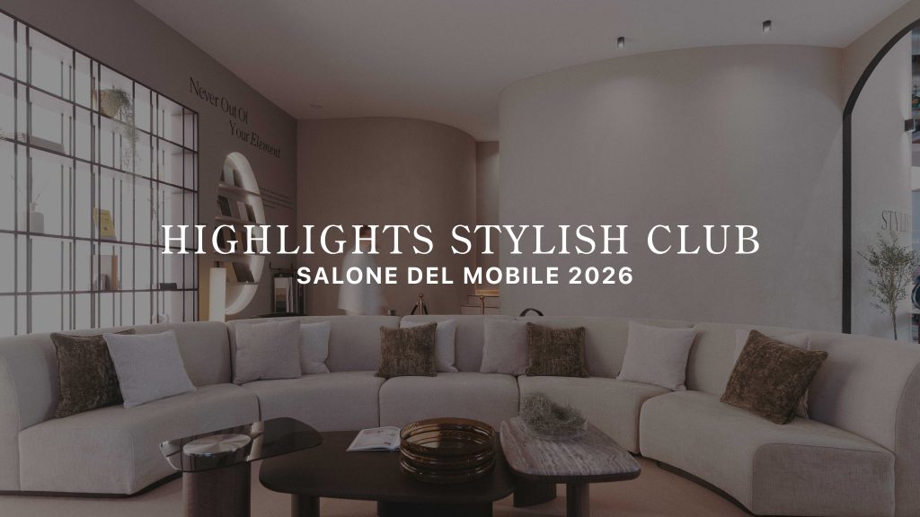 The living area features a curved sectional sofa layered with soft neutral cushions, paired with two sculptural tables styled with warm, tactile accents. A curved wall and open shelving frame the space, while a visitor passes by beneath the message “Never Out of Your Element,” reinforcing the stand’s calm, immersive atmosphere. This setting reflects refined living room furniture, shaped by the clarity of contemporary furniture, and a layout designed to spark subtle living room inspiration.