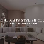 The living area features a curved sectional sofa layered with soft neutral cushions, paired with two sculptural tables styled with warm, tactile accents. A curved wall and open shelving frame the space, while a visitor passes by beneath the message “Never Out of Your Element,” reinforcing the stand’s calm, immersive atmosphere. This setting reflects refined living room furniture, shaped by the clarity of contemporary furniture, and a layout designed to spark subtle living room inspiration.