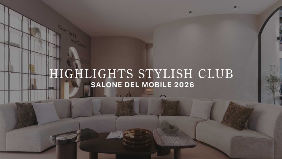 The living area features a curved sectional sofa layered with soft neutral cushions, paired with two sculptural tables styled with warm, tactile accents. A curved wall and open shelving frame the space, while a visitor passes by beneath the message “Never Out of Your Element,” reinforcing the stand’s calm, immersive atmosphere. This setting reflects refined living room furniture, shaped by the clarity of contemporary furniture, and a layout designed to spark subtle living room inspiration.