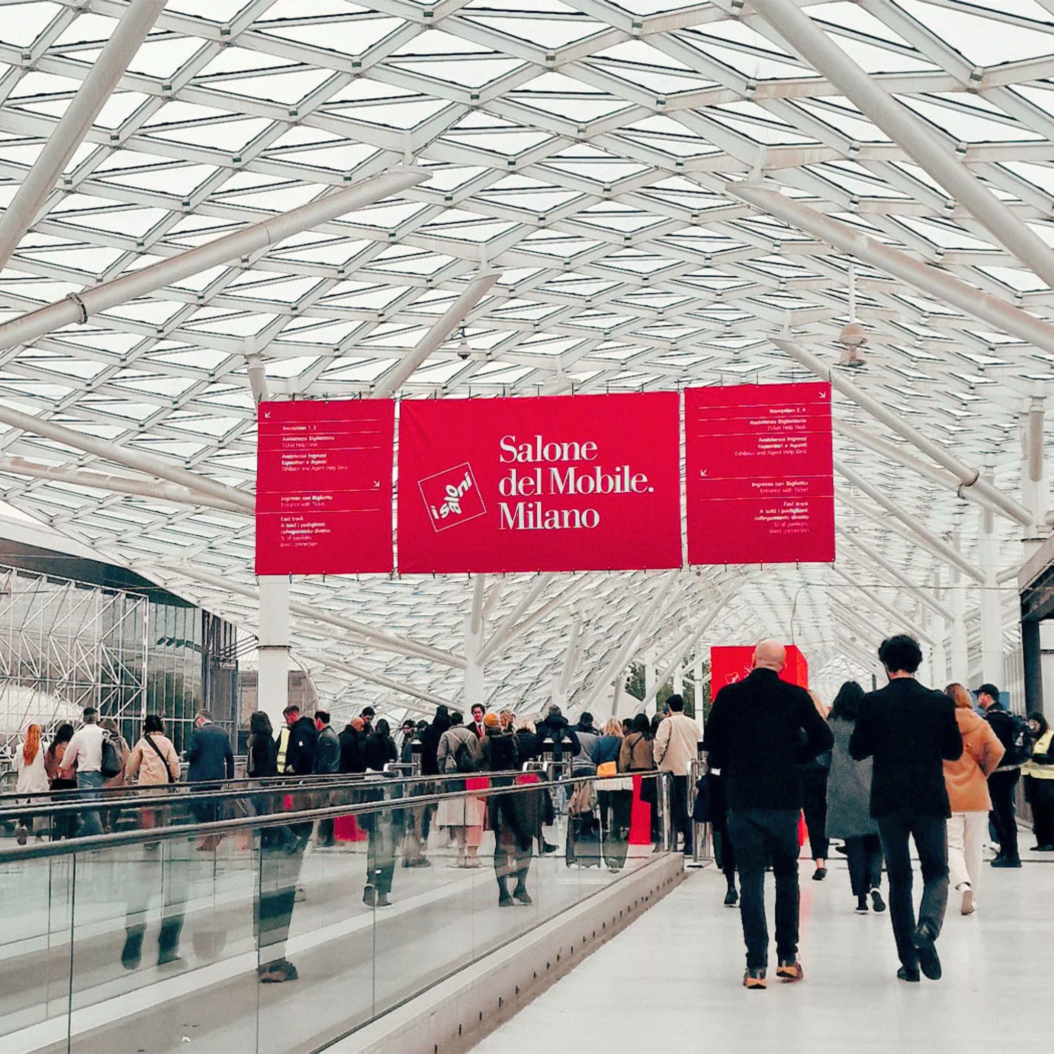 Crowds move through the central walkway at Fiera Milano Rho beneath the iconic red Salone del Mobile banner, illustrating the scale and energy of the fair. The architectural structure guides visitors through the exhibition halls, supporting the flow of furniture design exploration, helping newcomers navigate luxury furniture showcases, and framing the experience of contemporary design, furniture luxury design, and modern furniture at the event.