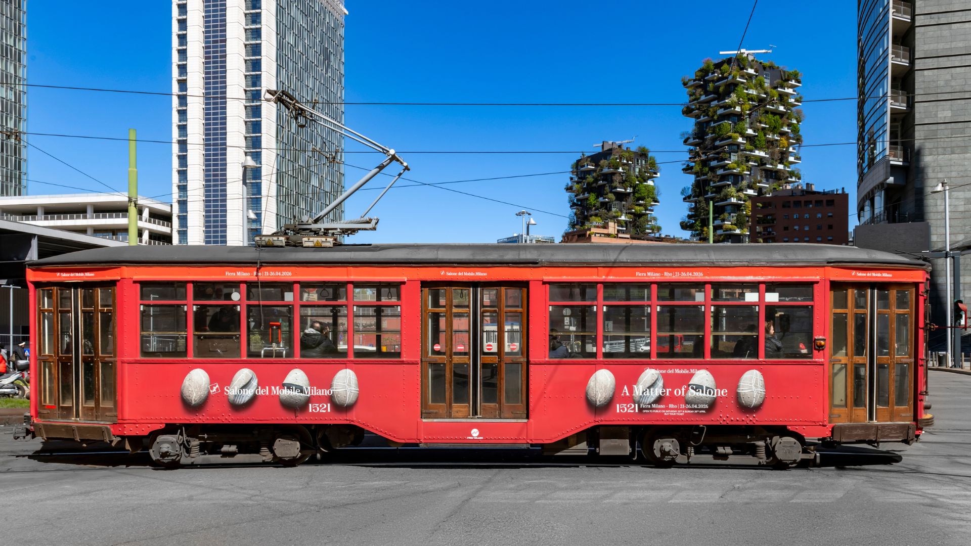 A red Milan tram featuring Salone del Mobile branding moves through the city, highlighting one of the transport options visitors encounter during Design Week. The scene reflects how Milan’s public network supports easy movement across the city, helping attendees navigate public transport, reach key design events, and travel efficiently during Milan Design Week, connecting the urban experience with interior design inspiration and the evolving world of modern furniture.