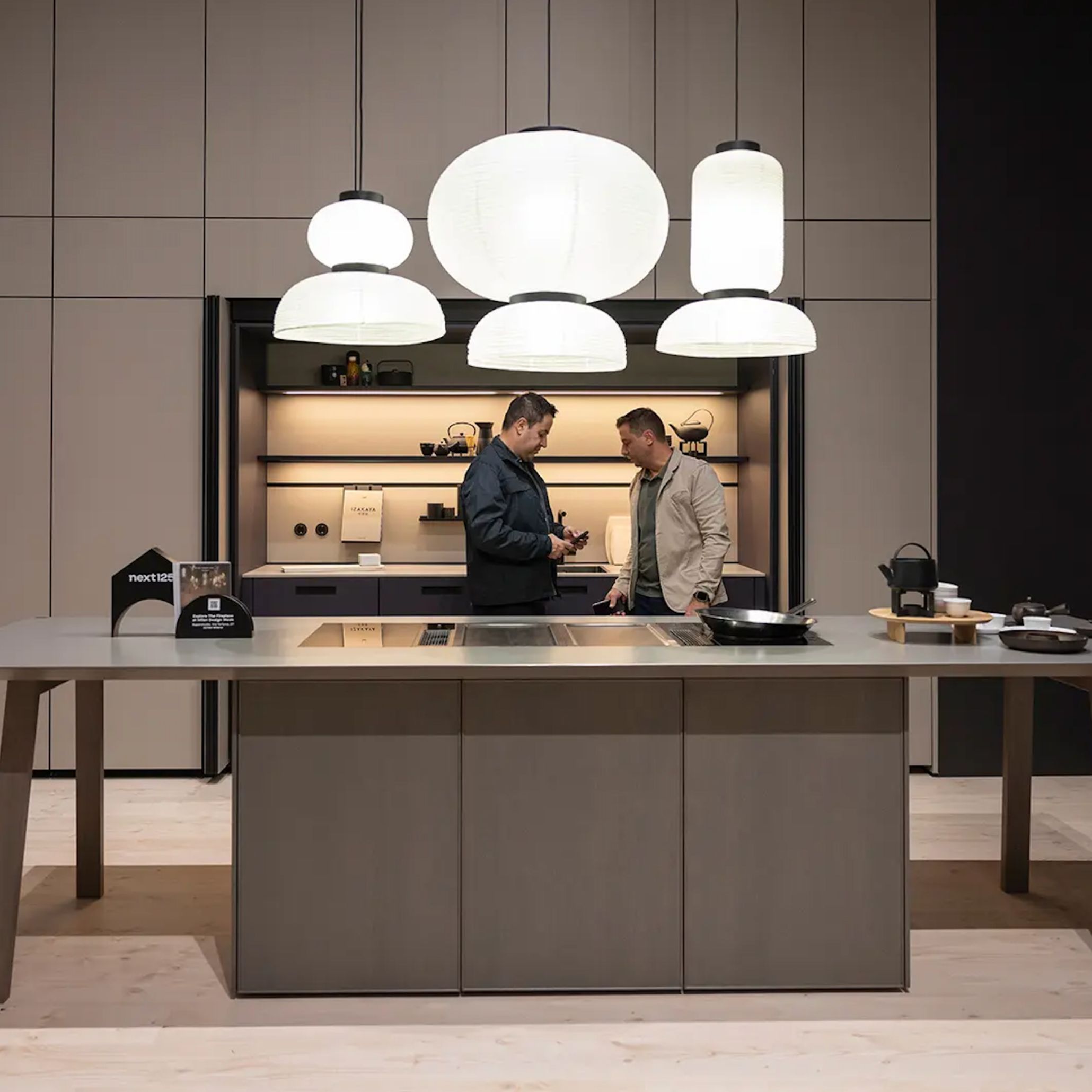 Visitors explore a contemporary kitchen display at EuroCucina, featuring a large island, integrated appliances, and warm lighting that highlights functional design. The scene reflects how modern kitchens prioritise efficient layouts and material quality, showcasing advances in kitchen design, the role of home technology, and evolving interior solutions connected to modern kitchen furniture and kitchen inspiration.