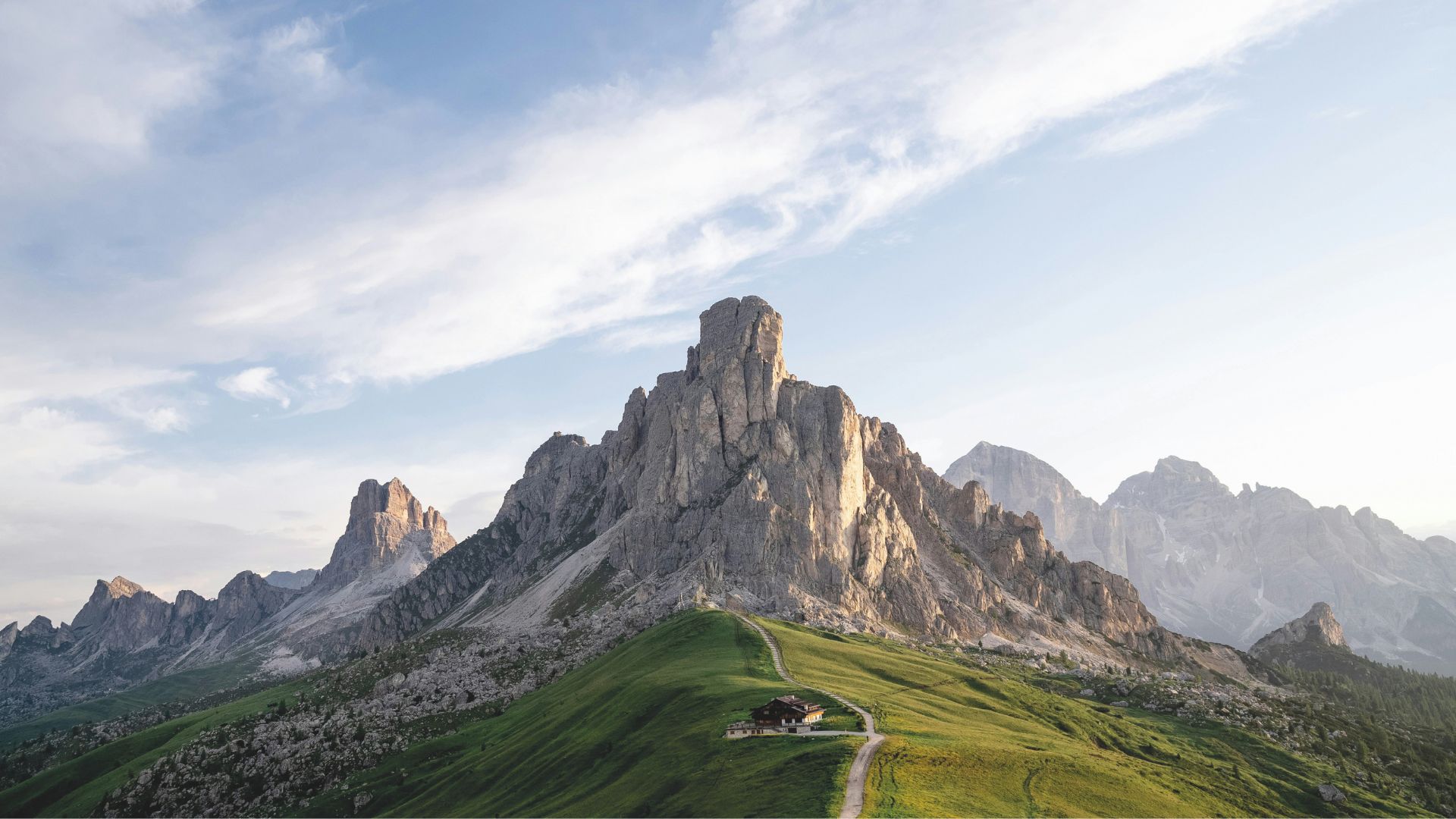 A dramatic mountain peak emerging from a green hillside, with a small lodge anchored at its base and a winding path leading upward. Warm light grazes the rocky cliffs, revealing the raw textures and quiet strength of the landscape. The scene evokes the grounding presence of natural materials, the serene balance found in minimalist design, and the timeless harmony that inspires interior design inspiration.
