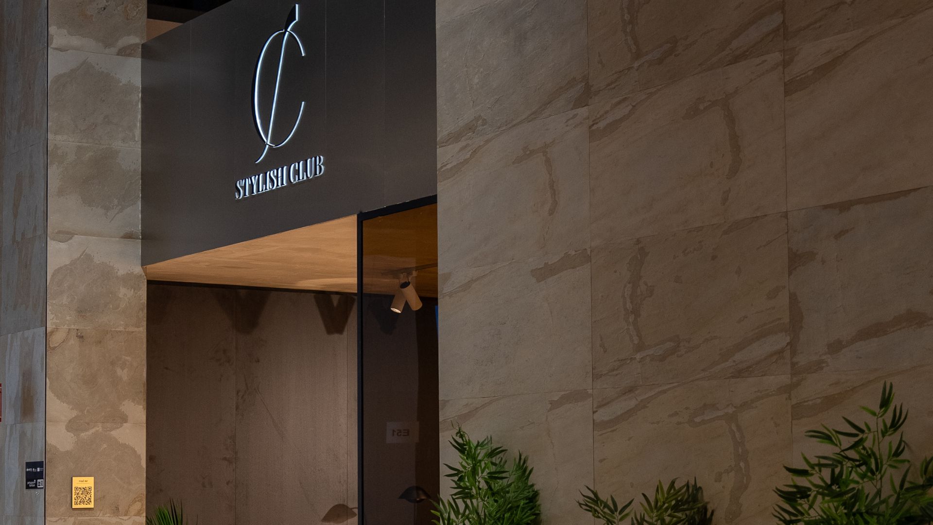 A refined entrance to the Stylish Club stand, framed by warm stone walls and illuminated signage featuring the brand’s logo. Soft lighting and greenery introduce a sense of calm, while the clean lines and natural materials reflect the brand’s commitment to modern design, minimalist architecture, and the crafted elegance of luxury furniture. The composition evokes a quiet, intentional atmosphere, offering interior design inspiration rooted in balance and material purity.