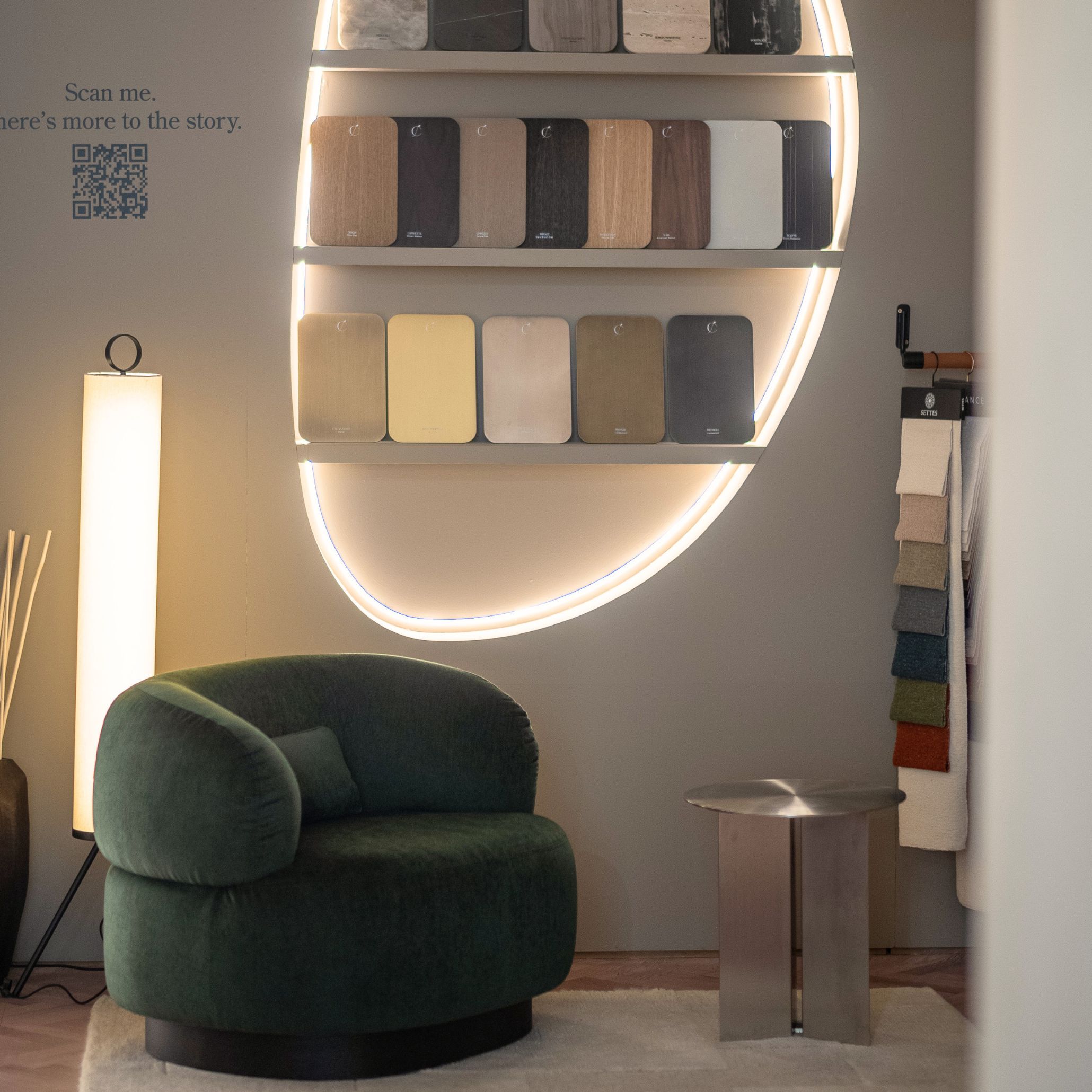 The reading corner presents a quiet, intimate atmosphere where the Dolomites Armchair sits beside a minimalist side table, framed by a glowing display of material samples and soft ambient lighting. The curved wall and suspended fabric swatches highlight the tactile richness of the collections, offering a moment to pause and explore textures up close. This serene setting reflects the brand’s approach to sophisticated bedroom furniture, complemented by the crafted presence of contemporary furniture and a layout designed to spark subtle interior design inspiration.