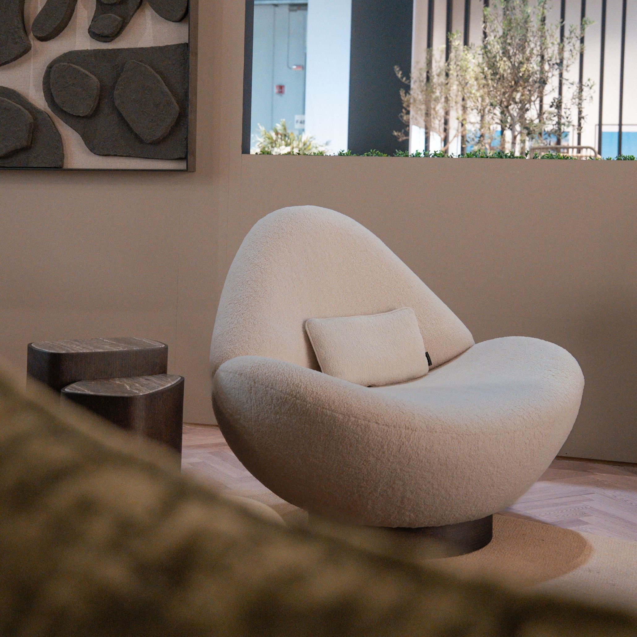A sculptural Stone Armchair anchors this intimate corner, complemented by the Dolomites Side Table and framed by organic artwork and natural light. The soft, rounded forms and tactile materials create a serene moment within the stand, inviting visitors to slow down and observe the details. The composition highlights the elegance of luxury home furniture, balanced with the crafted identity of bespoke furniture and a quiet sense of living room inspiration.