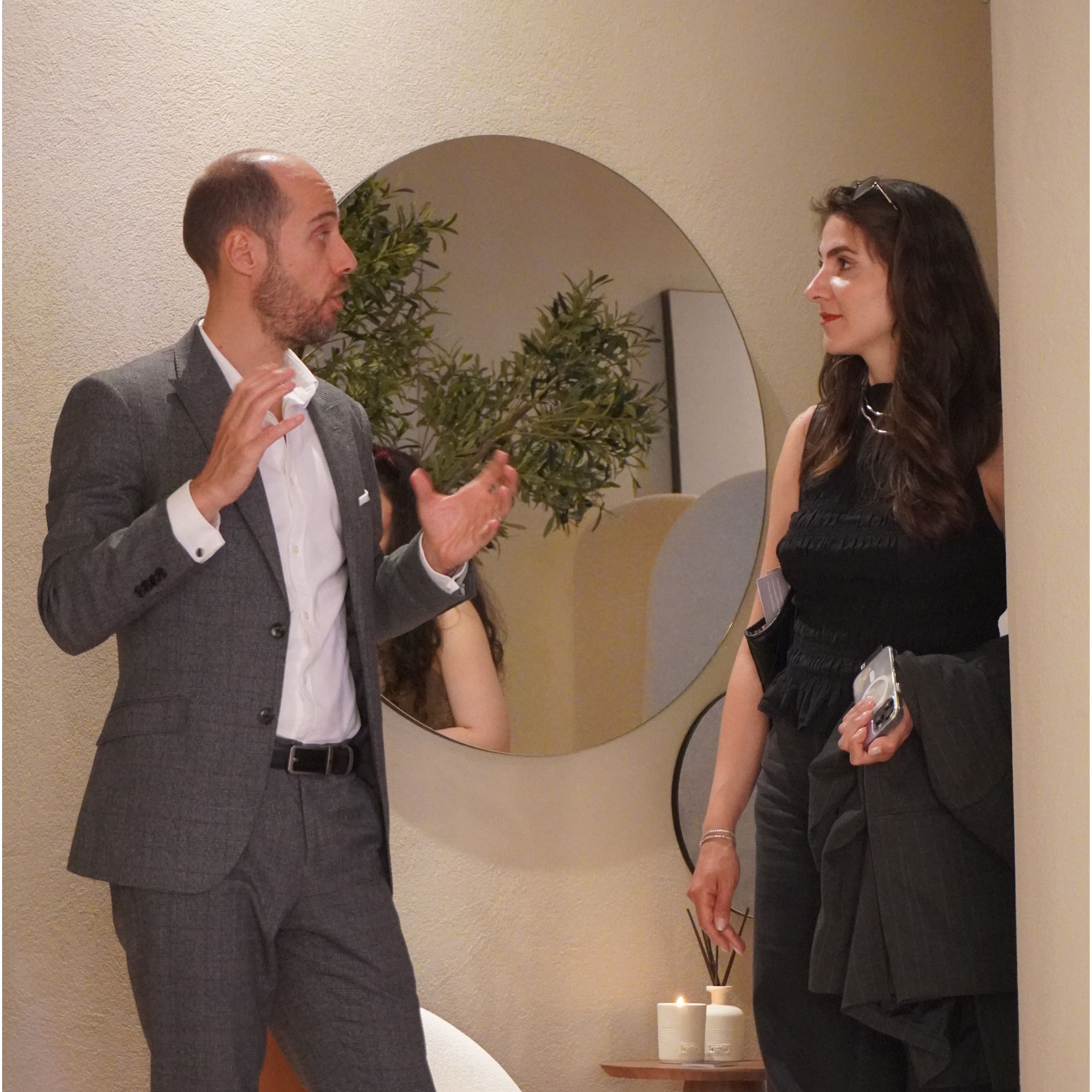 A Stylish Club team member speaks with a client in a warm, softly illuminated setting, surrounded by natural textures and subtle decorative elements. A circular mirror reflects greenery and candlelight, reinforcing the intimate and welcoming mood that shaped many encounters throughout the fair. This moment captures the human side of hospitality furniture, the inviting presence of luxury furniture, and the atmosphere of shared interior design inspiration that defined Salone del Mobile 2026.