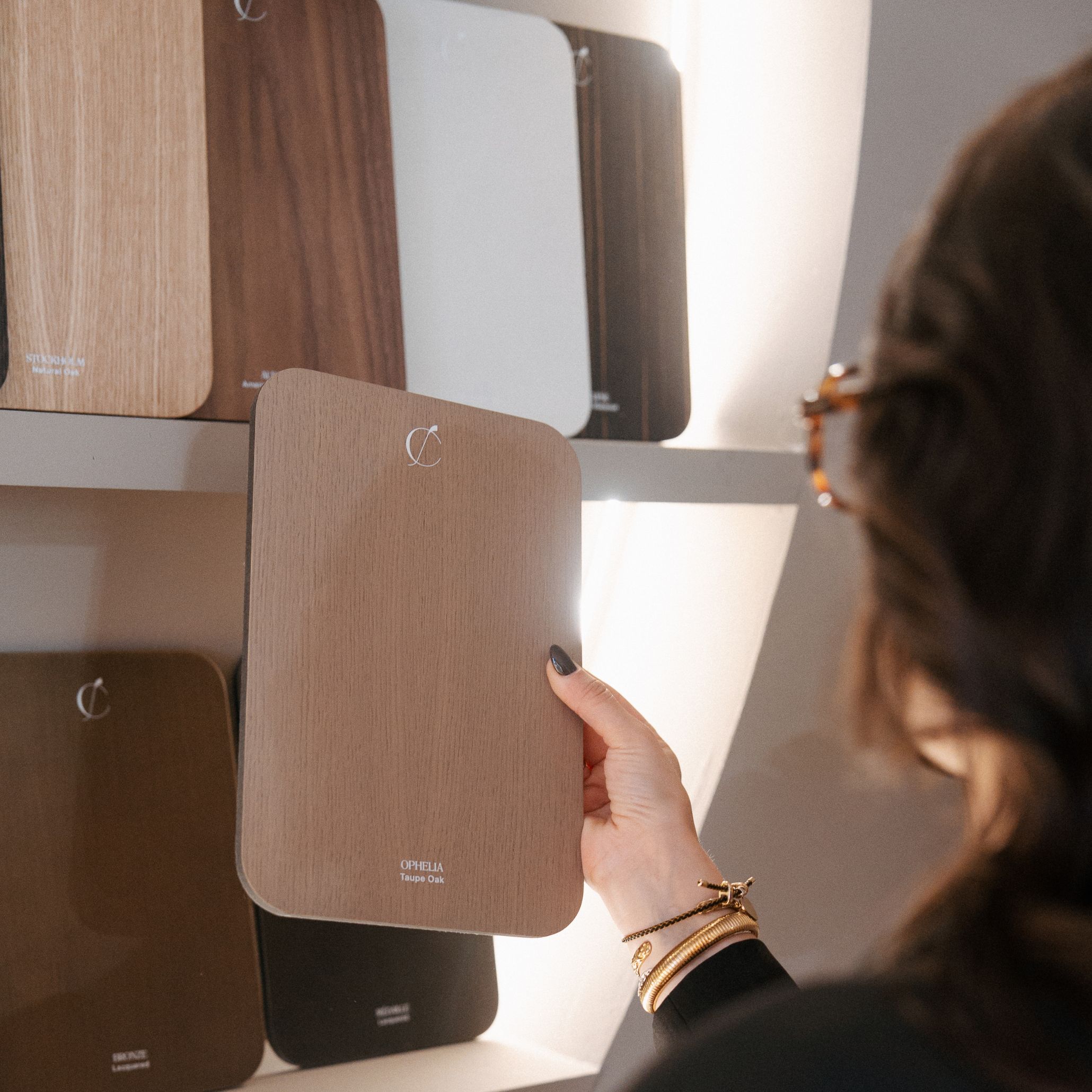 A visitor holds a wood sample from the material display wall, surrounded by an array of finishes that highlight the brand’s commitment to texture, craftsmanship, and natural inspiration. The illuminated shelving showcases a curated palette of woods and colors, offering a tactile introduction to the collections. This display reflects the precision of custom furniture, the richness of finest wood, and the thoughtful curation that supports meaningful design details.
