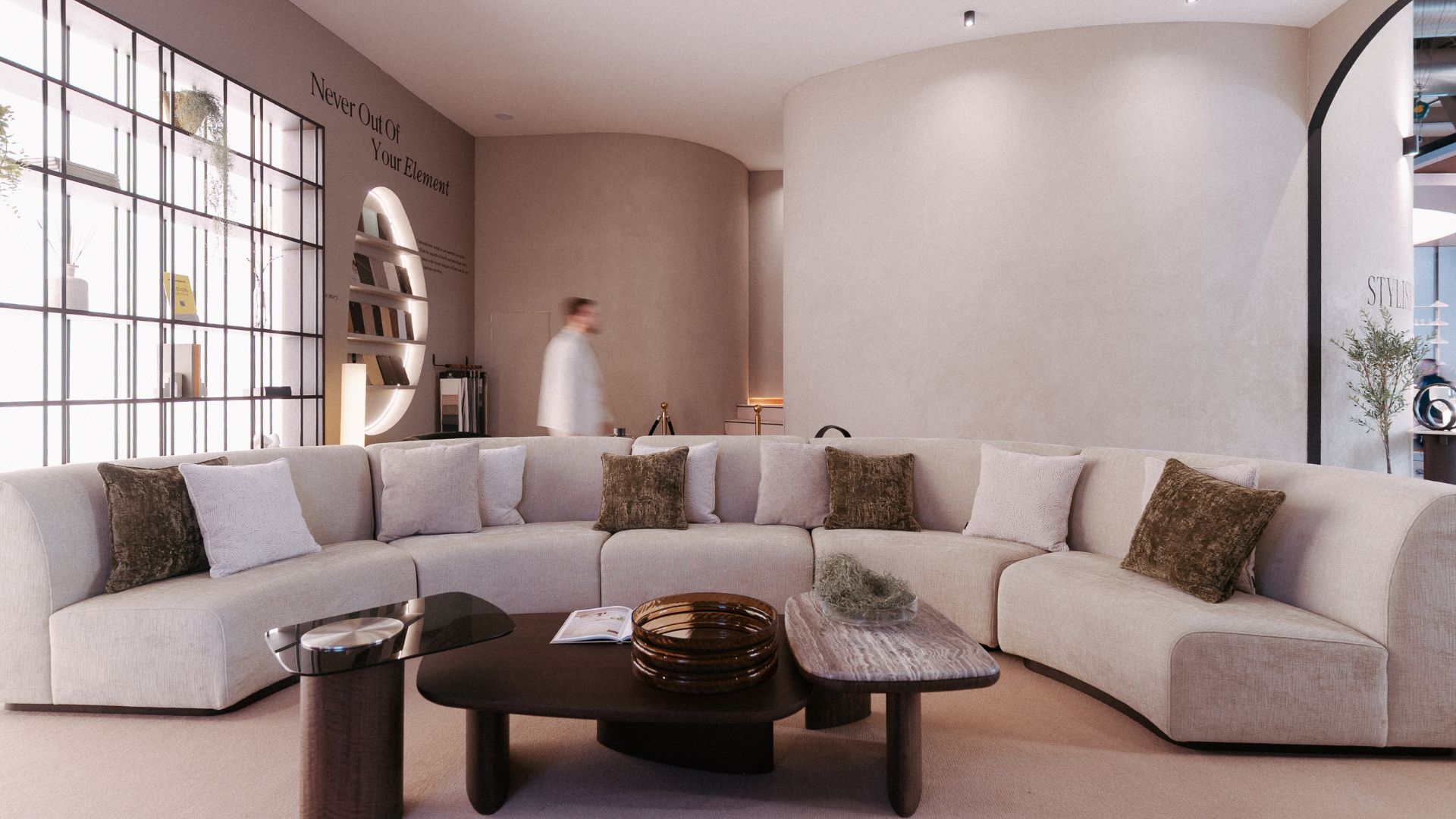 The Stone Sofa anchors the living area with soft, curved lines, paired with the Dolomites Coffee Table styled with warm, natural accents. A curved wall and open shelving frame the scene as a visitor walks by, adding quiet movement to the space. The composition reflects refined living room furniture, shaped by the clarity of modern furniture and a calm sense of living room inspiration.