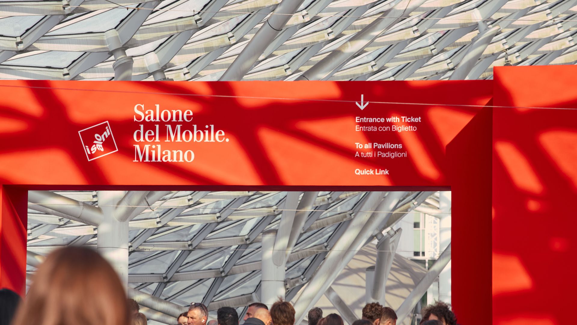 Red entrance structure for Salone del Mobile 2026 stands against a geometric glass-and-steel canopy as visitors walk toward the pavilions, capturing the energy of Milan during design week. The scene reflects the movement of people gathering to explore new directions in interiors, shaped by modern furniture, enriched by interior design inspiration, and connected to the evolving world of luxury furniture highlighted throughout the fair.