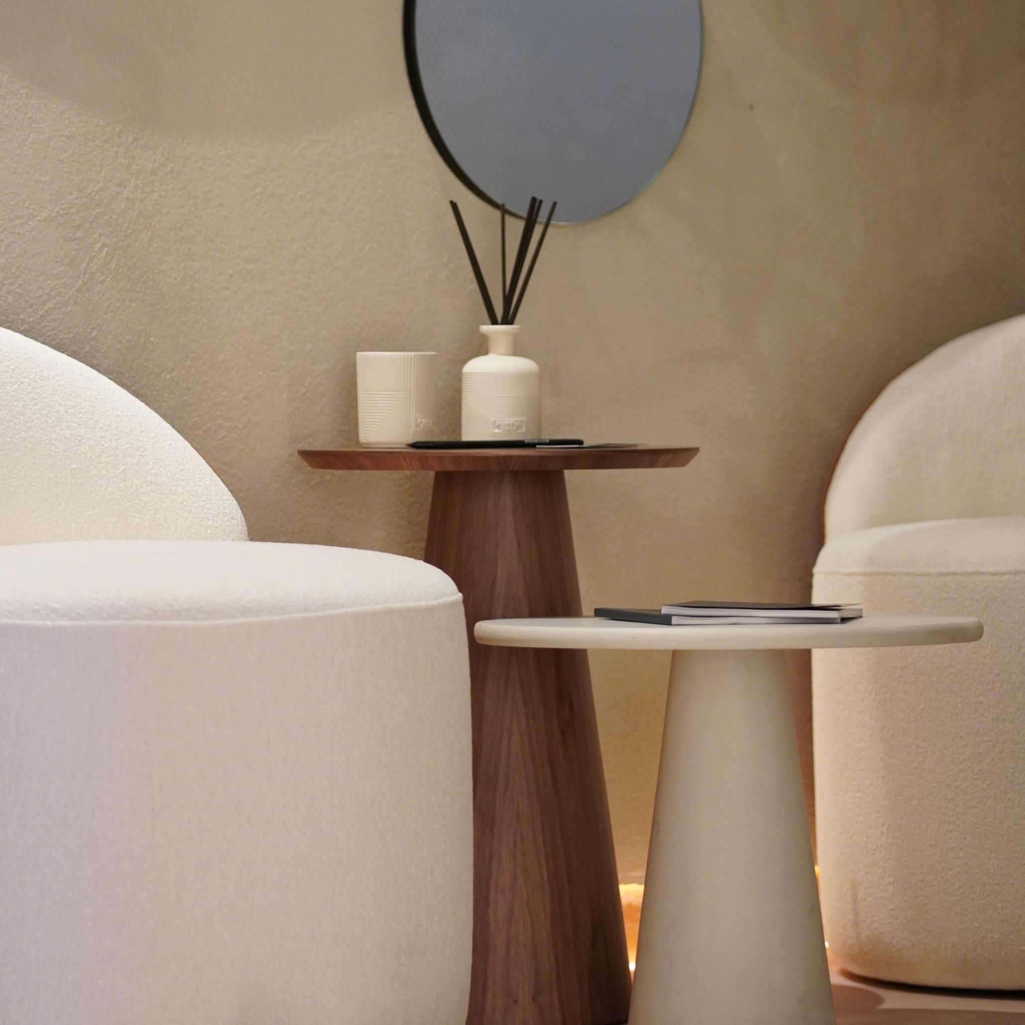 The Olivier Side Table in microconcrete sits between two rounded cream stools, paired with a wooden side table holding a diffuser and ceramic cup. The soft lighting and textured wall create a calm, tactile setting shaped by living room furniture, enriched by luxury home furniture, and connected to natural design details.