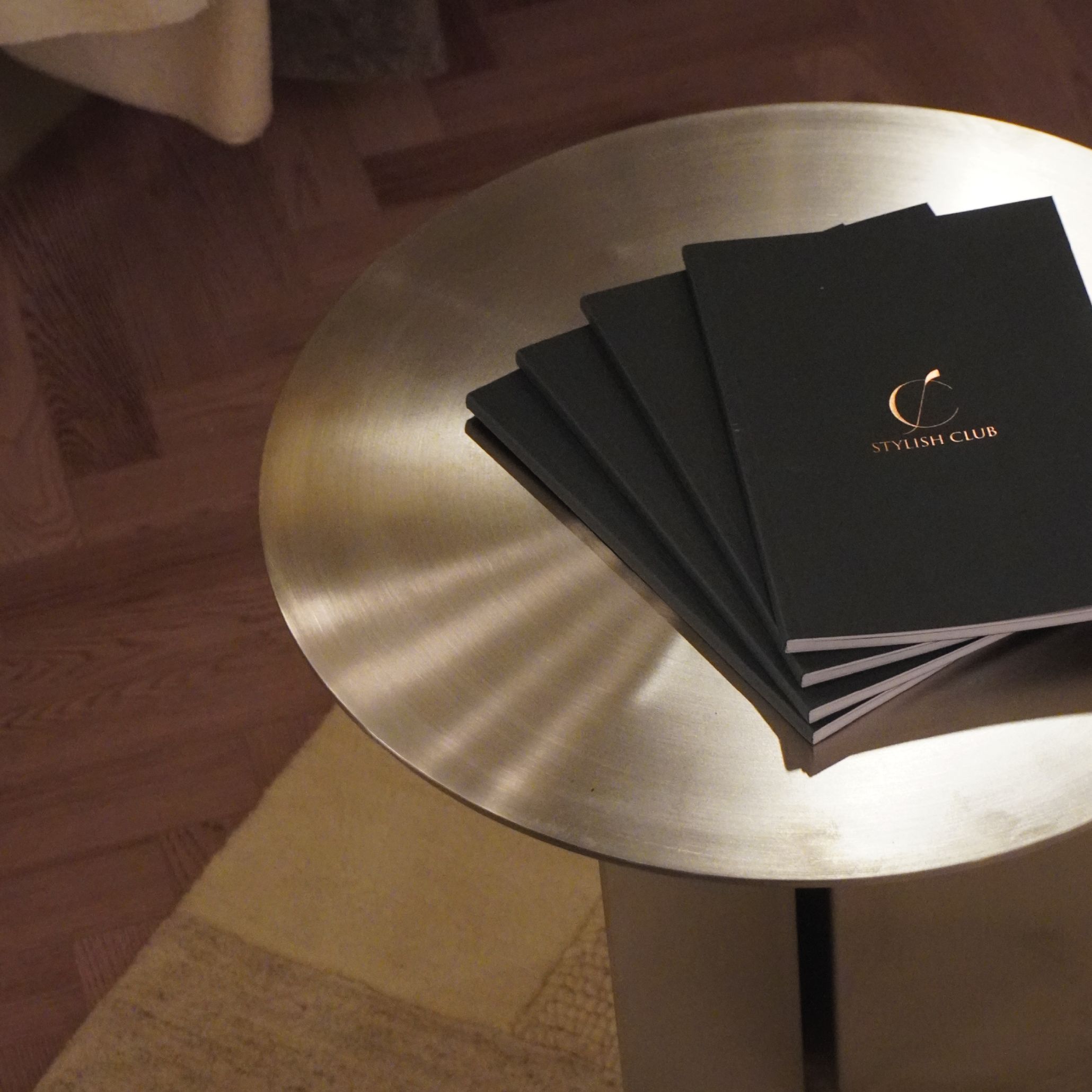 A brushed Steel Side Table displays a fan‑shaped arrangement of Stylish Club brochures with gold accents, set on a wooden floor with a beige rug. The cool metallic finish reflects the trend toward expressive materials within modern furniture, complemented by refined luxury furniture, and aligned with contemporary interior design inspiration.