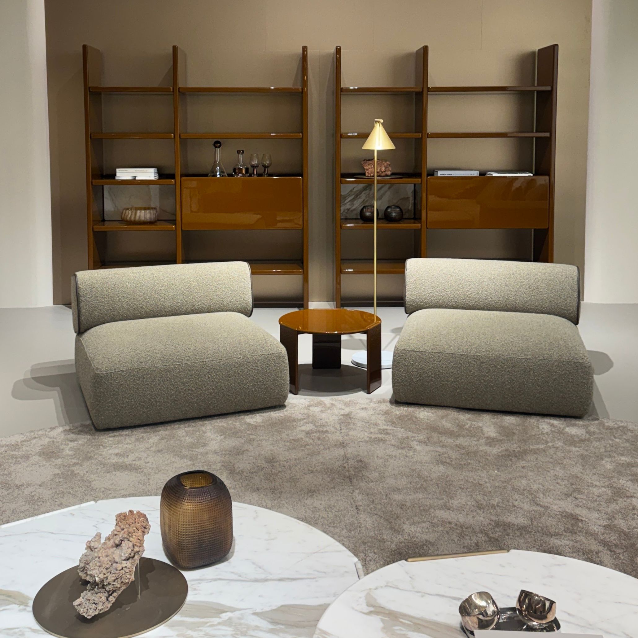 A pair of white marble coffee tables with sculptural decor sit in front of two low neutral lounge chairs, framed by tall brown shelving units and a soft beige carpet. The warm palette of browns, creams, and muted greys reflects Salone del Mobile’s shift toward enveloping, grounded interiors shaped by living room furniture, enriched by luxury home furniture, and guided by atmospheric interior design inspiration.