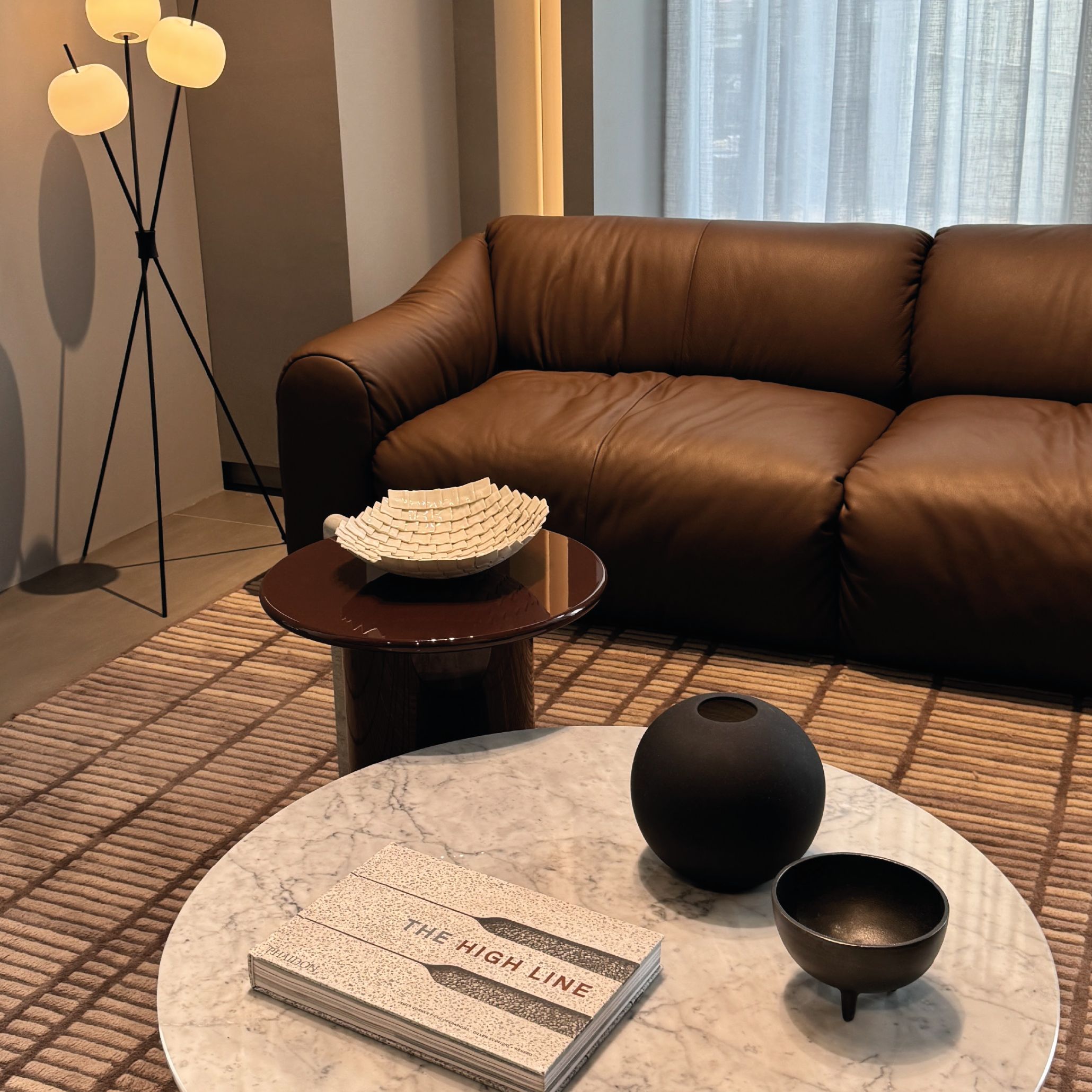 A brown leather sofa faces a marble coffee table styled with black bowls and a design book, paired with a small side table and a floor lamp with frosted globes. The warm, earthy tones and soft lighting echo the 2026 move toward intimate, expressive palettes within modern furniture, complemented by luxury furniture, and connected to refined design details.