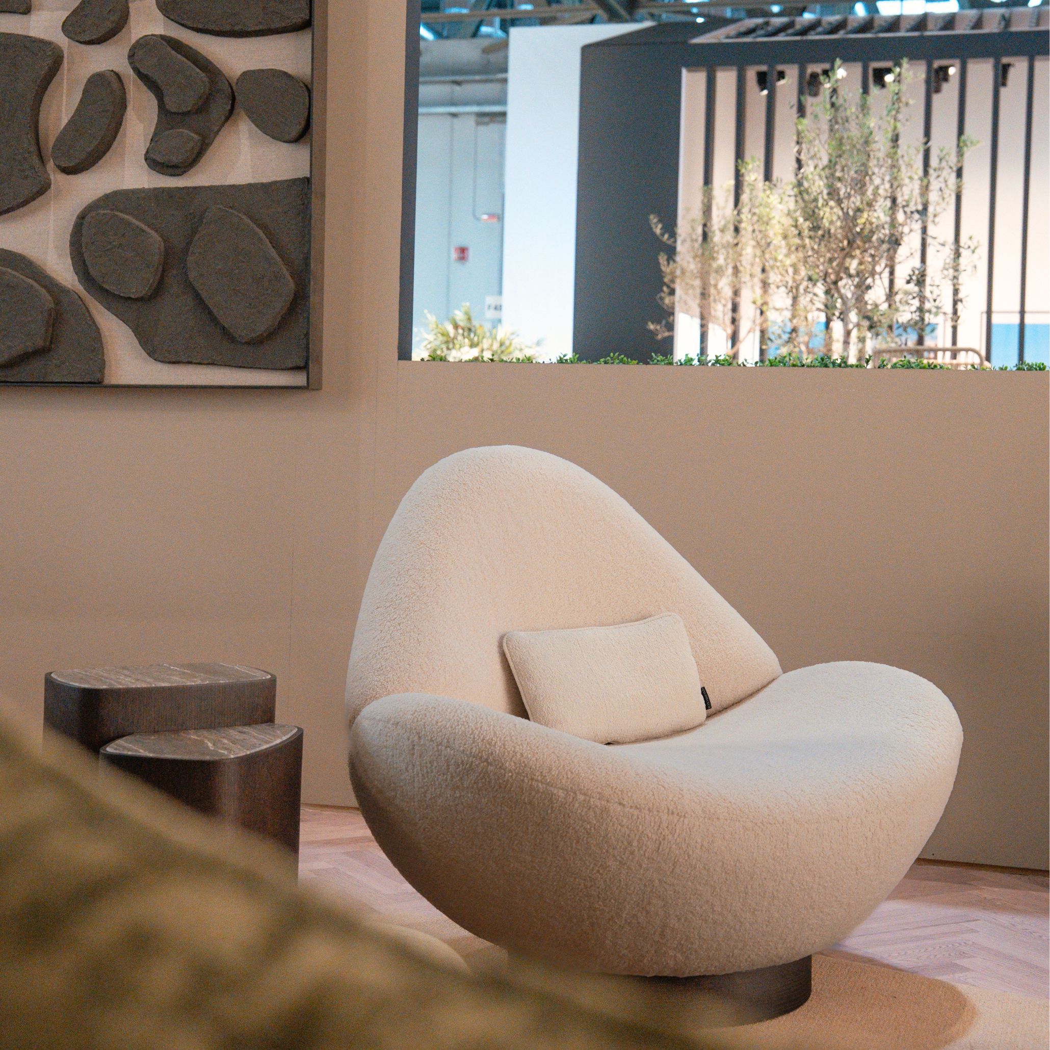 A cream, organic Stone armchair with a soft cushion sits besides a dark wooden side table, framed by an abstract stone‑textured wall piece and a view toward an outdoor area with vertical slats and greenery. The fluid silhouettes and natural textures highlight the rise of organic volumes within luxury home furniture, shaped by living room furniture, and enriched by tactile design details.
