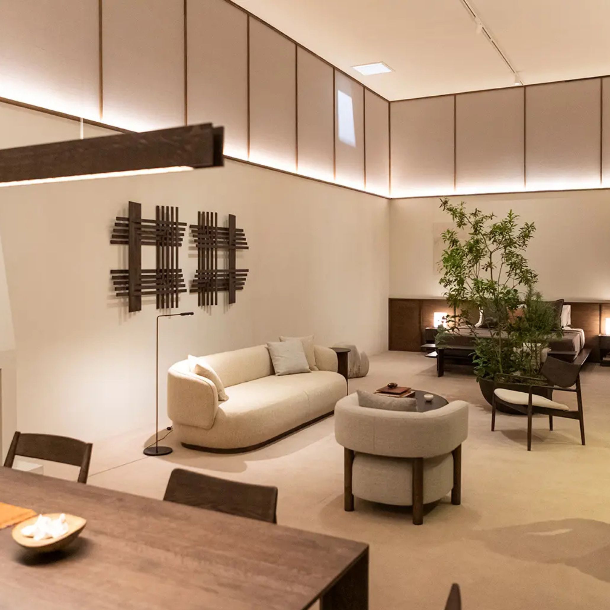 A beige curved sofa and matching armchair anchor a warm, minimalist living space with a wooden coffee table, sculptural wall art, and a dining set in the foreground. The softened lines and rounded transitions reflect the 2026 shift toward human‑centred forms in modern furniture, complemented by luxury furniture, and connected to atmospheric interior design inspiration.