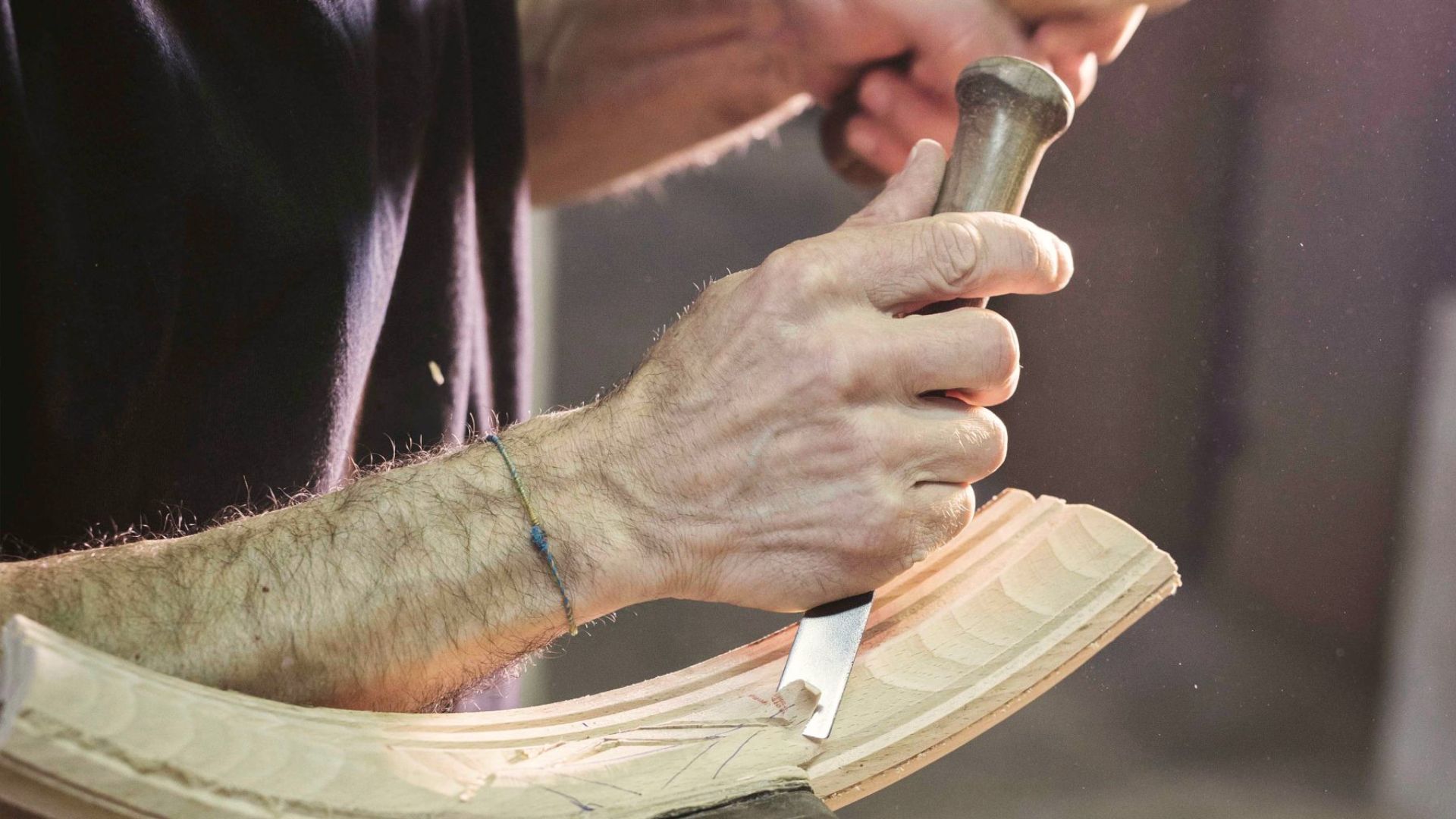 Hands carve a curved wooden surface with a chisel, revealing fine grooves and natural texture in the light-toned wood. The close-up highlights the human touch at the core of Salone del Mobile 2026, where expressive materials and intentional making shape custom furniture, elevate luxury handmade furniture, and reinforce the value of authentic design details.